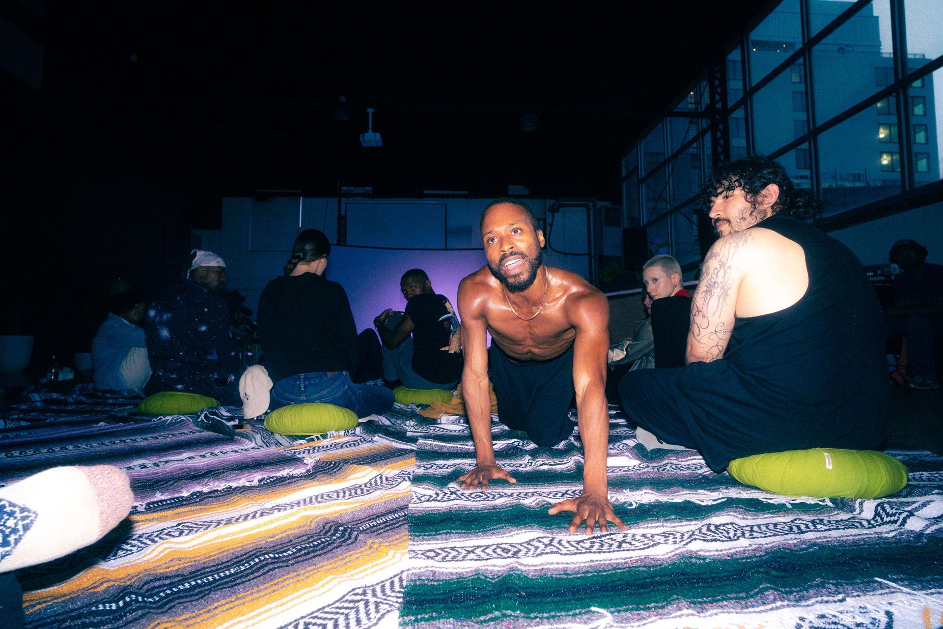 Group yoga class in a dark room. A shirtless Black man does push-ups, smiles. Others sit on cushions.