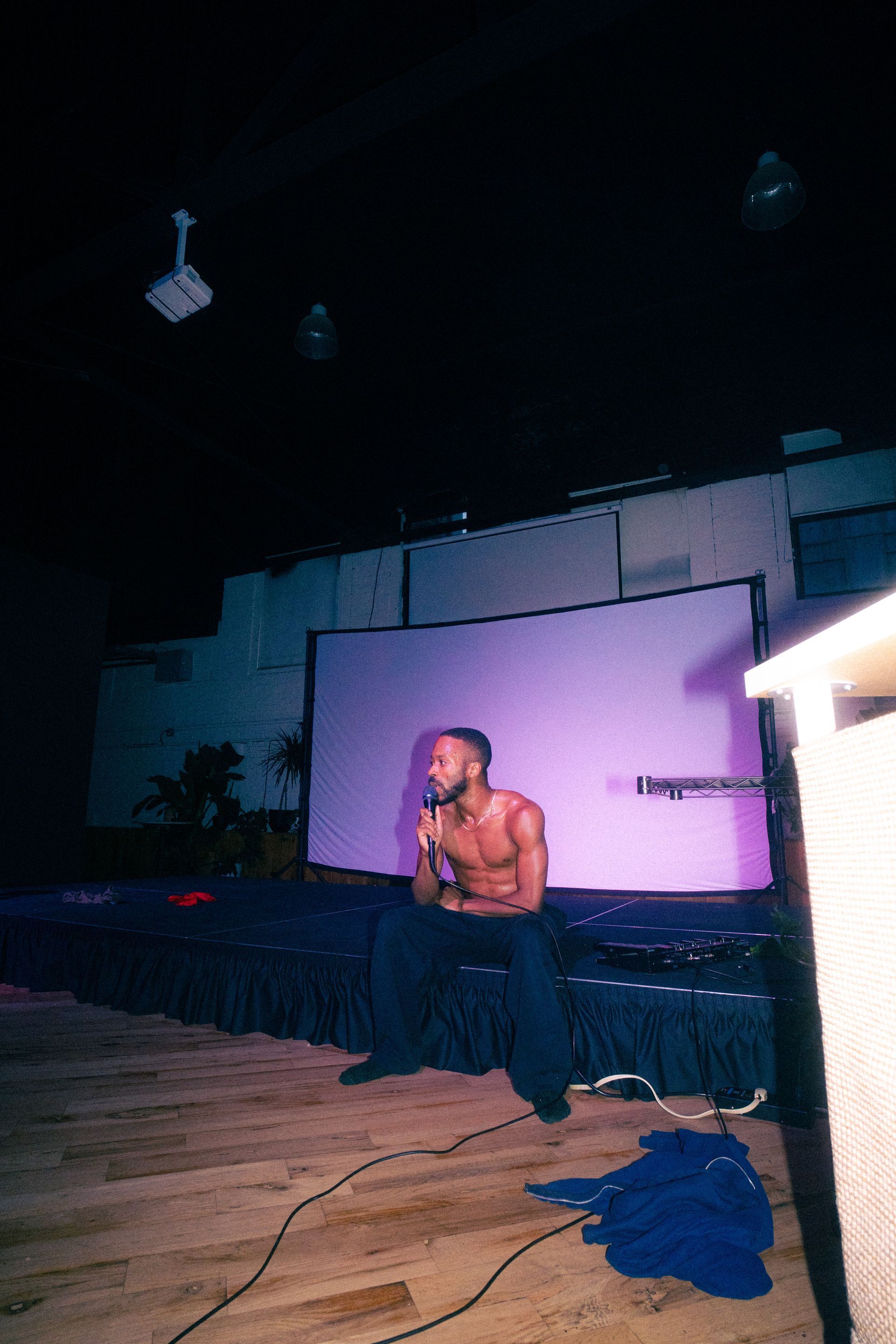 A shirtless Black man sits onstage in front of a projection screen. Dim purple lighting.