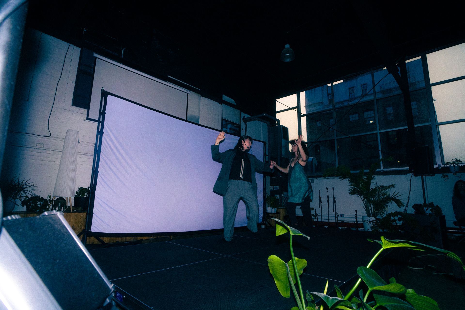 Two people dancing in front of a screen at an outdoor event, plants in the foreground, nighttime.