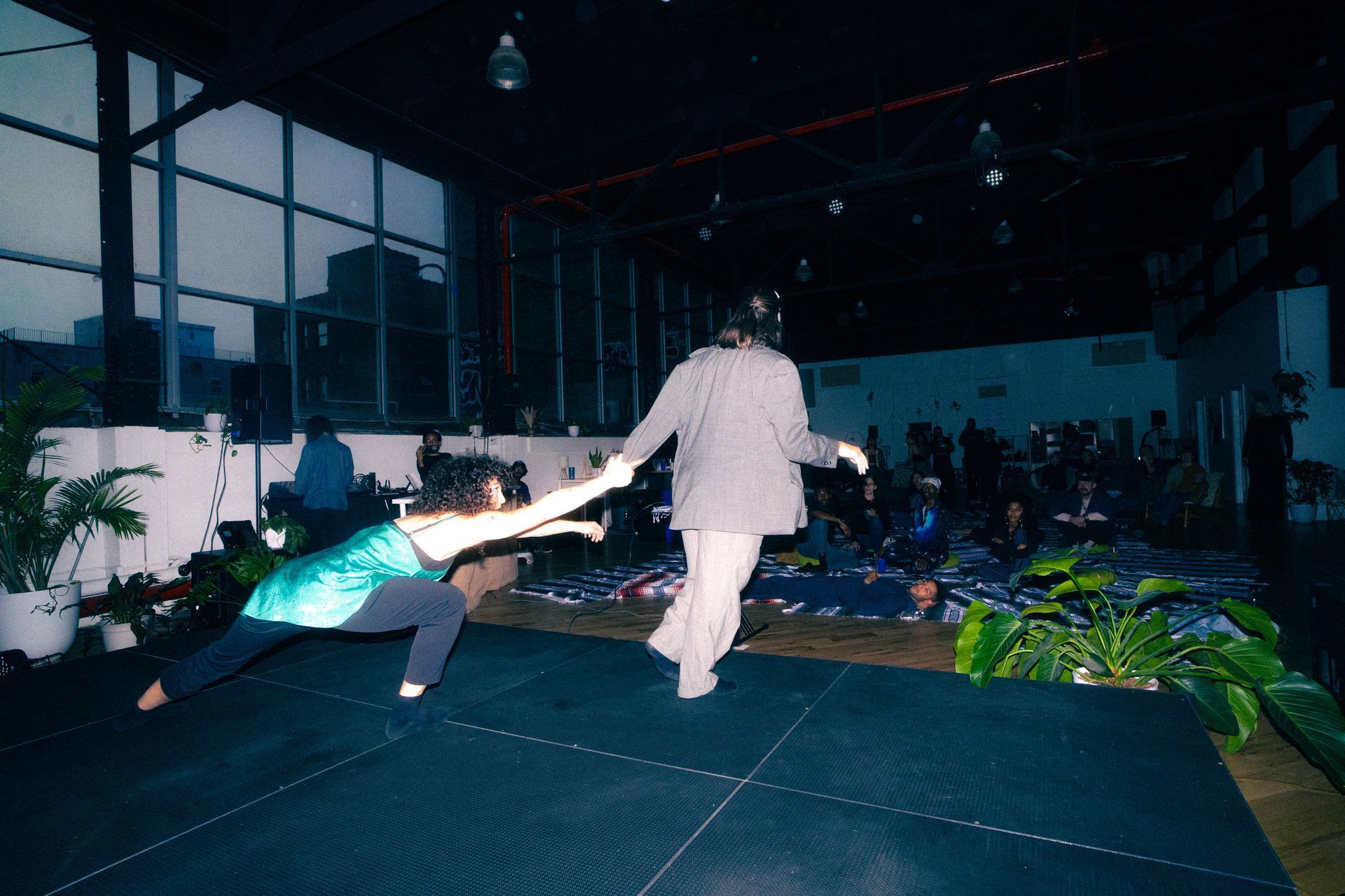 Two people dancing on stage; one reaching out, the other with arms outstretched. Dark room, audience seated.
