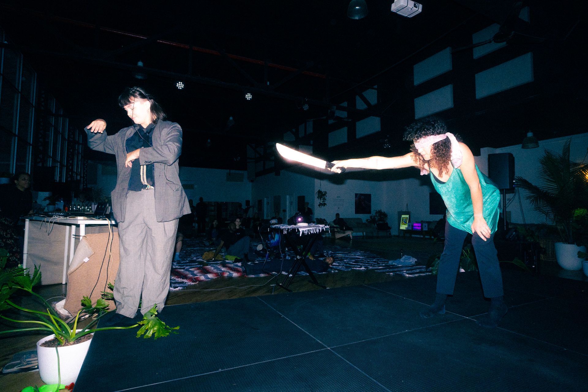Two people on a stage. One in a suit gestures, the other lunges forward holding a long, silver object. Dimly lit.
