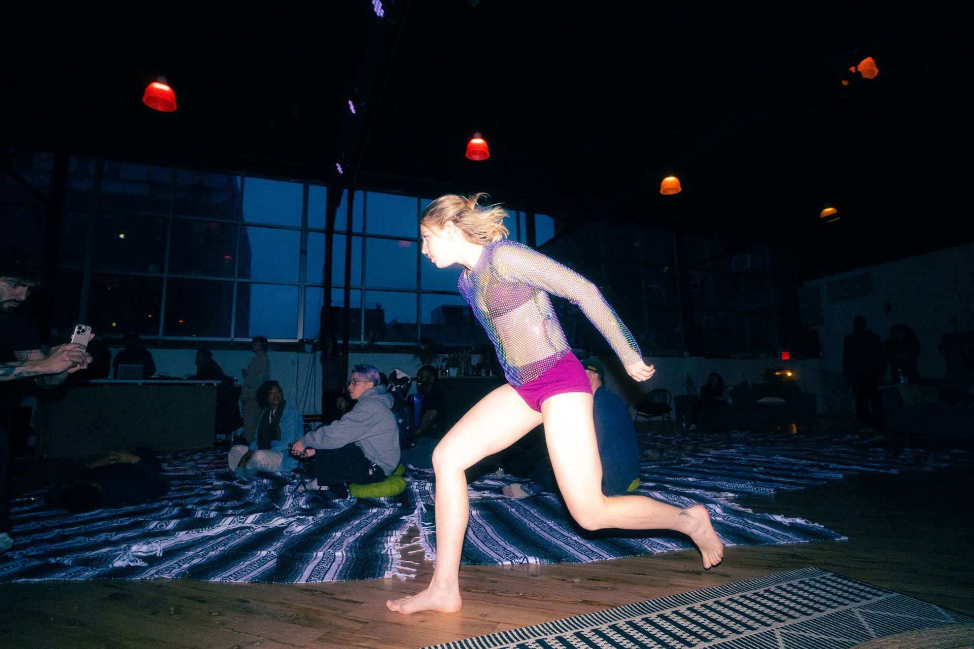 A person in a purple leotard and sheer top running, indoors, in front of a seated audience.
