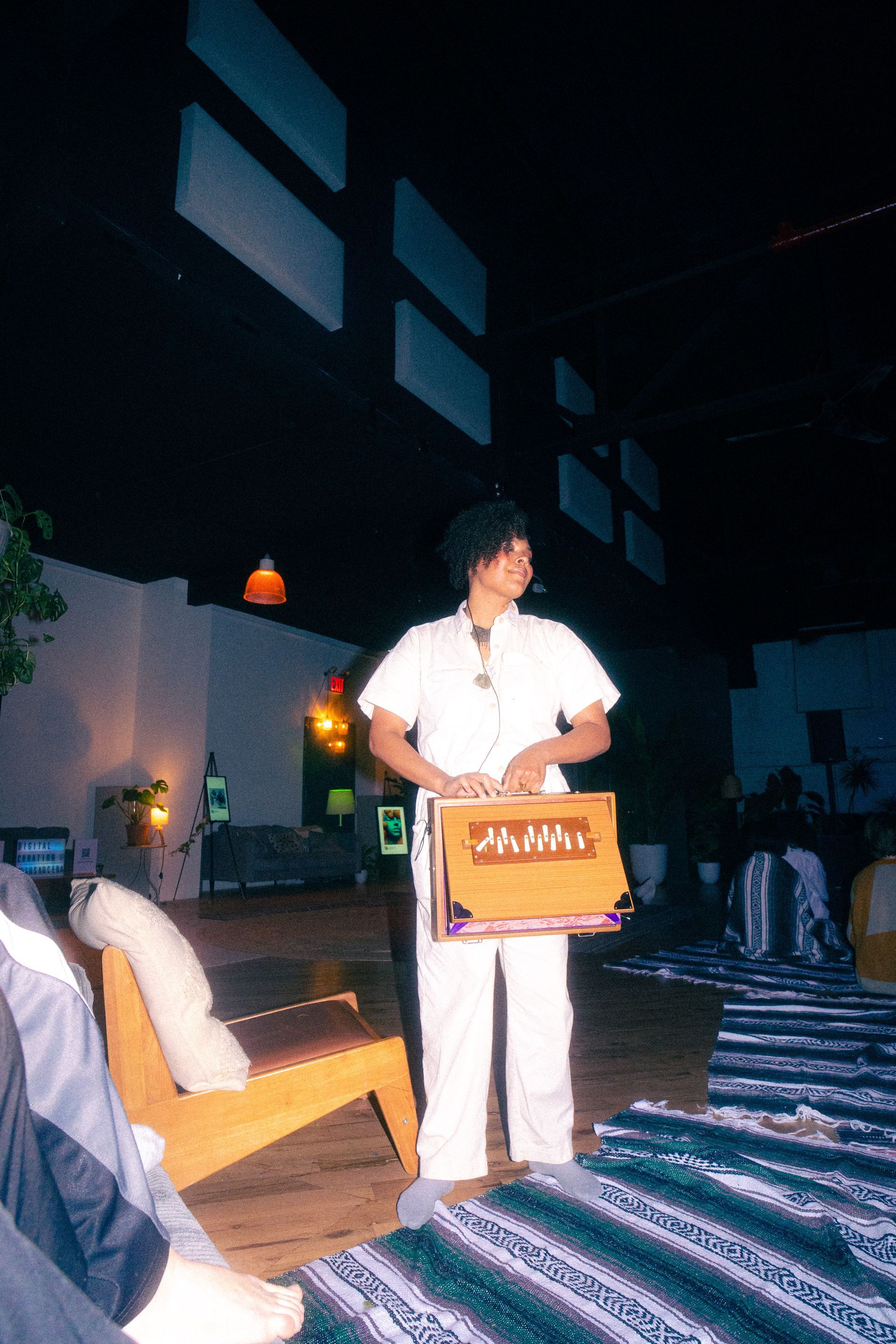 Person in white jumpsuit holding a brown box with gold lettering; standing on a rug. Interior setting.