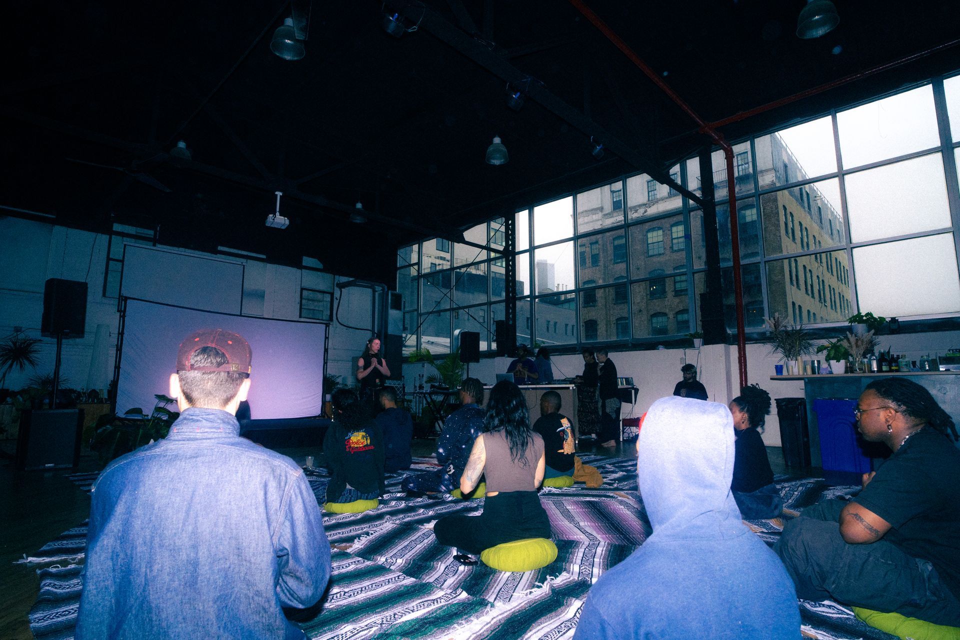 People seated on cushions, listening to a speaker in a large room with windows and an urban backdrop.