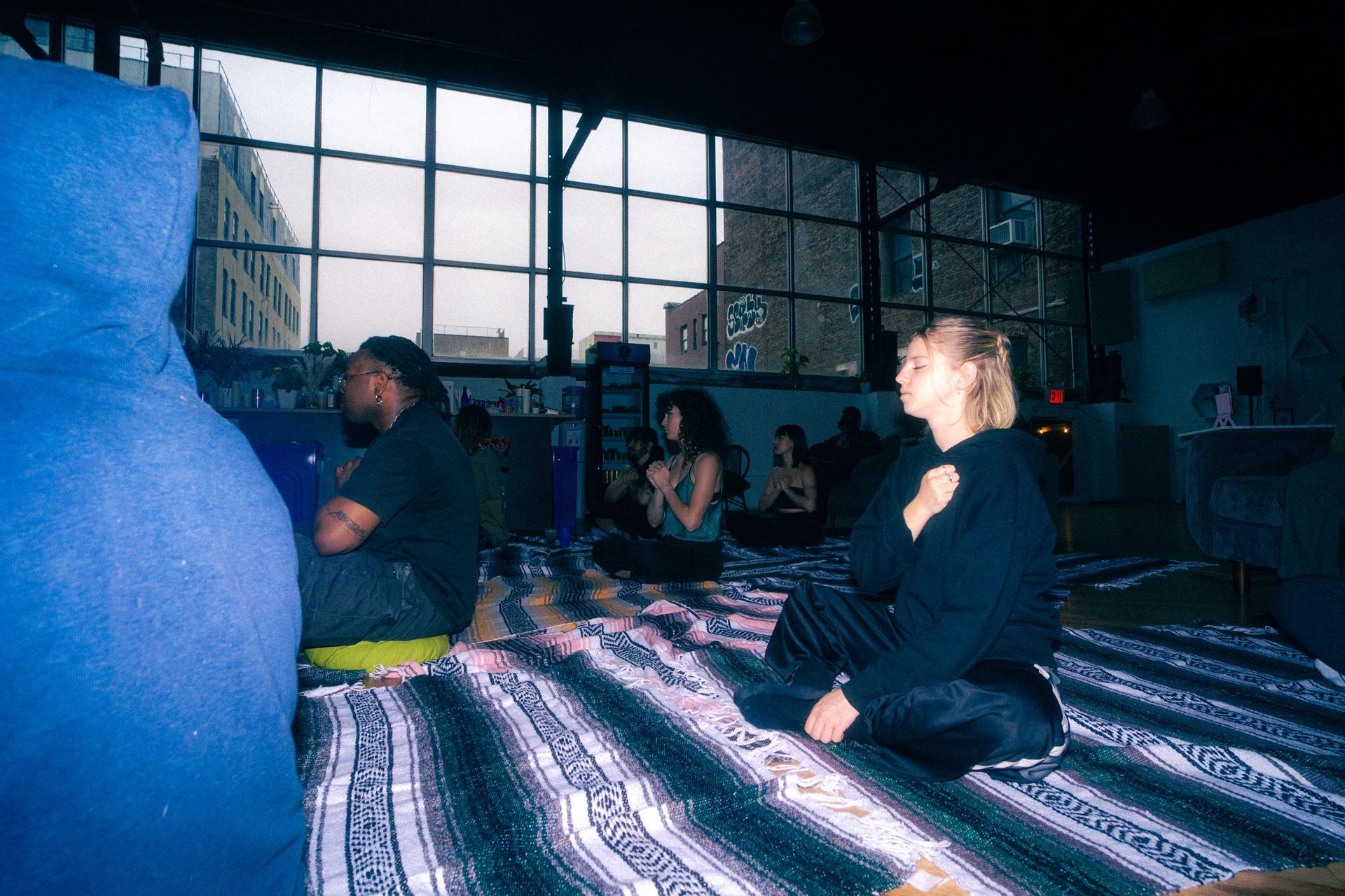 People in a dimly lit room sit on blankets, possibly meditating. Large windows are in the background.