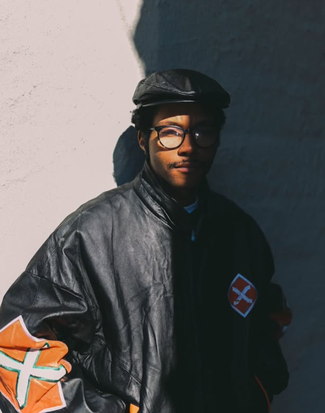 Man with glasses and newsboy cap in a leather jacket, standing outside in the sun.