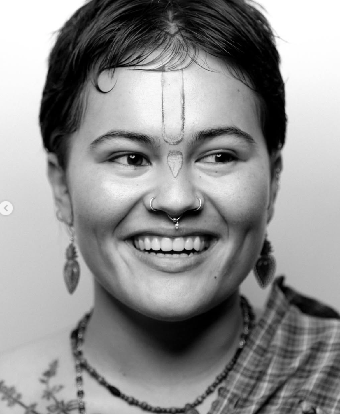 Smiling person with nose ring, ear ornaments, and forehead markings.