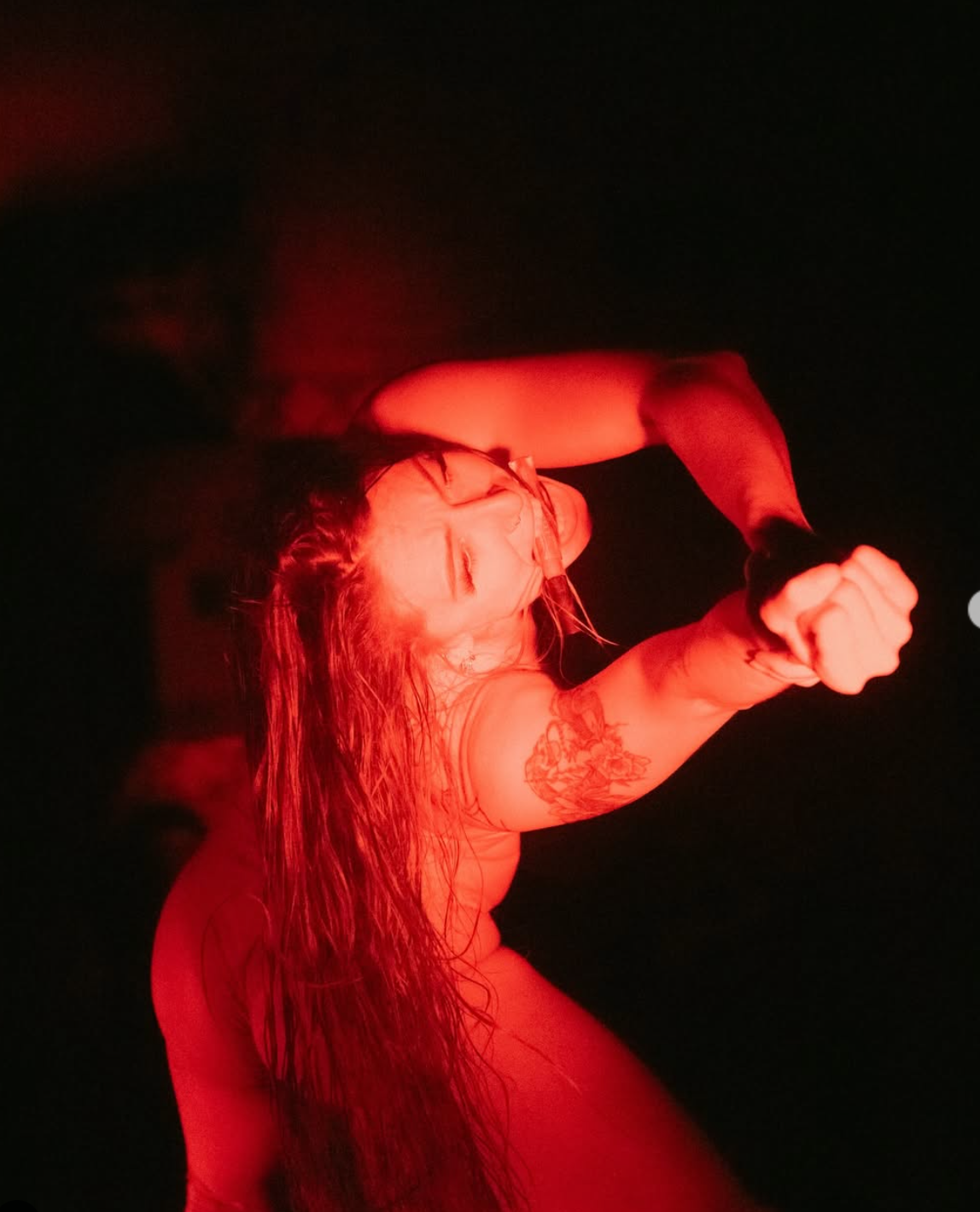 Woman in red light, long hair, arms raised, fist clenched, eyes closed, dark background.