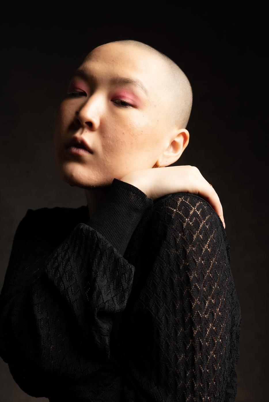 A bald person with pink eyeshadow, wearing a black top, poses in front of a dark background.