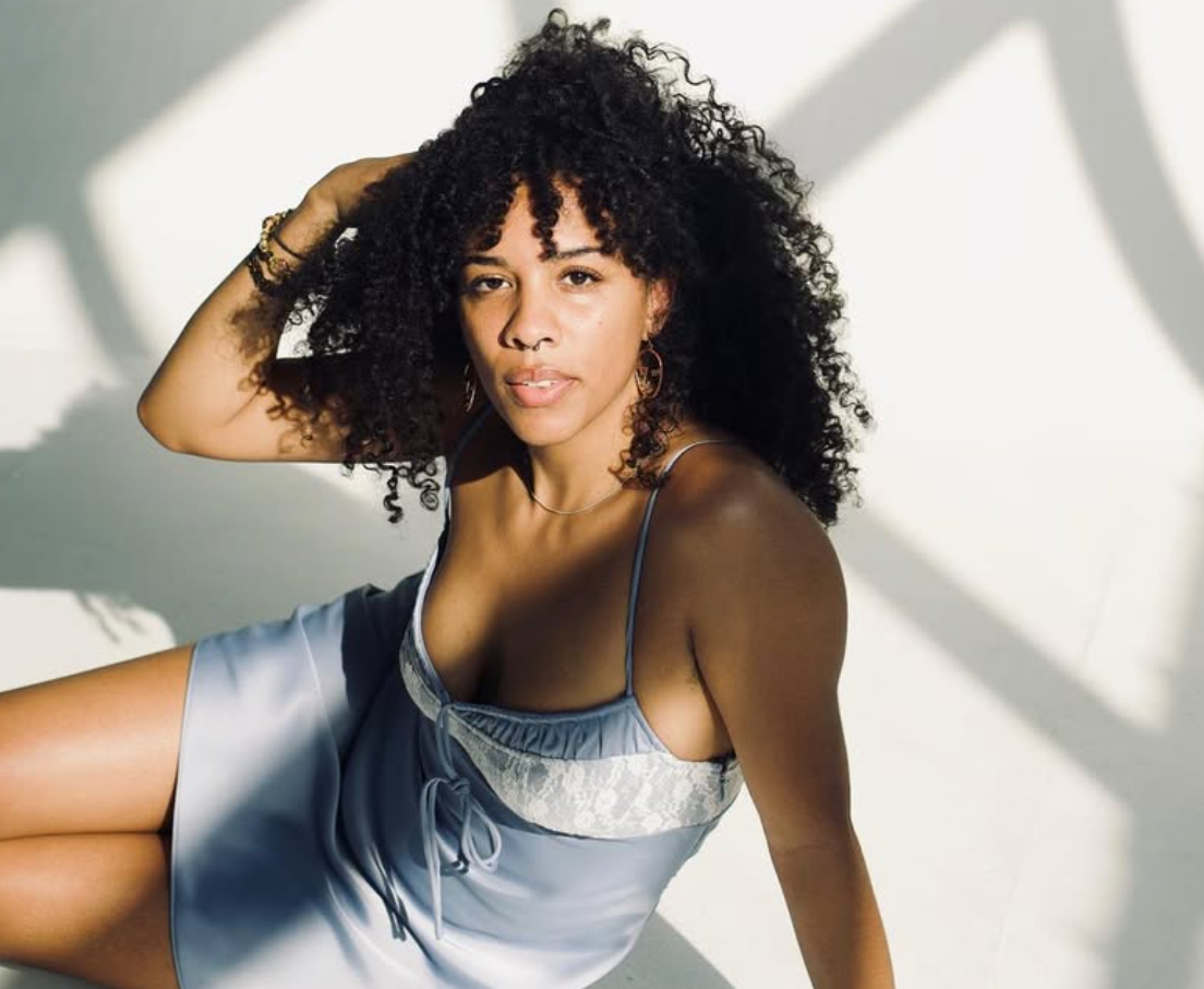 Woman with curly hair in a light blue dress, sitting with arm up, looking at the camera.