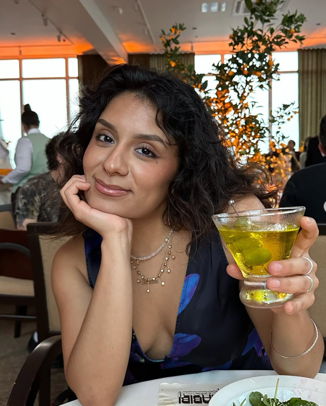 Woman in blue floral top holding a martini, resting chin on hand, in a restaurant.