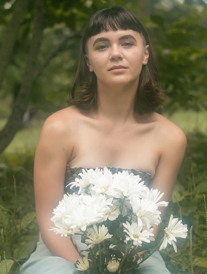 Woman holding white flowers, wearing strapless top, in a green outdoor setting.