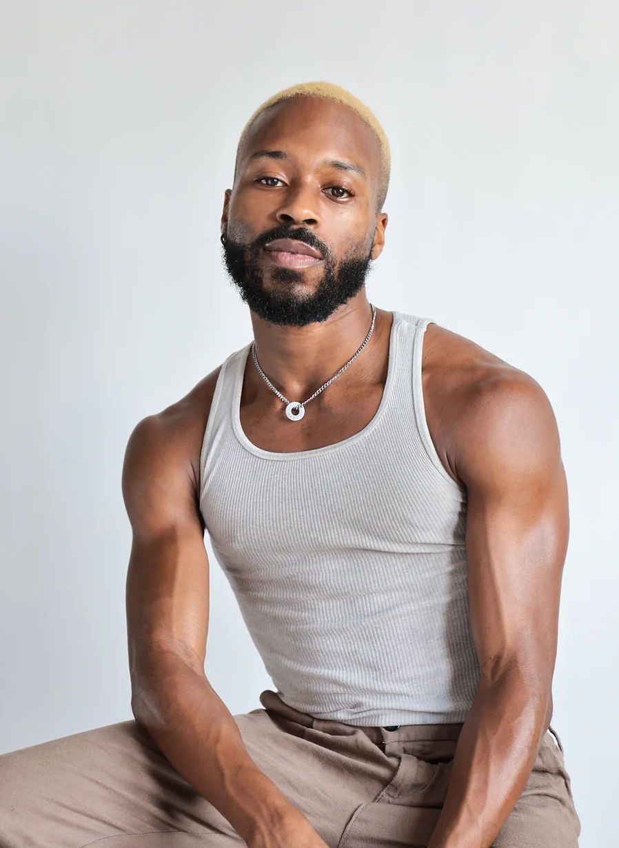 A Black man with blonde hair and a beard, wearing a white tank top and tan pants, poses seated, looking at the camera.