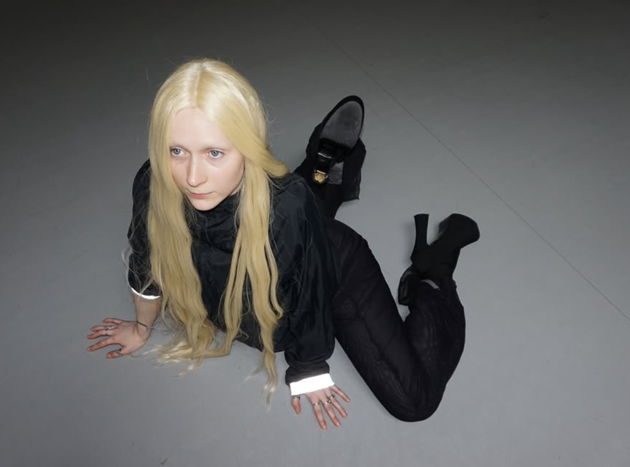 Blonde person in black outfit kneels on a grey floor, looking up.