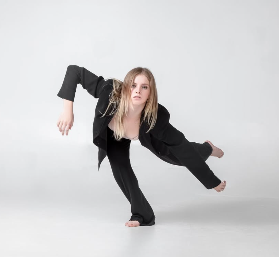 Blonde dancer in black suit balances, one leg extended, arm outstretched, studio setting.