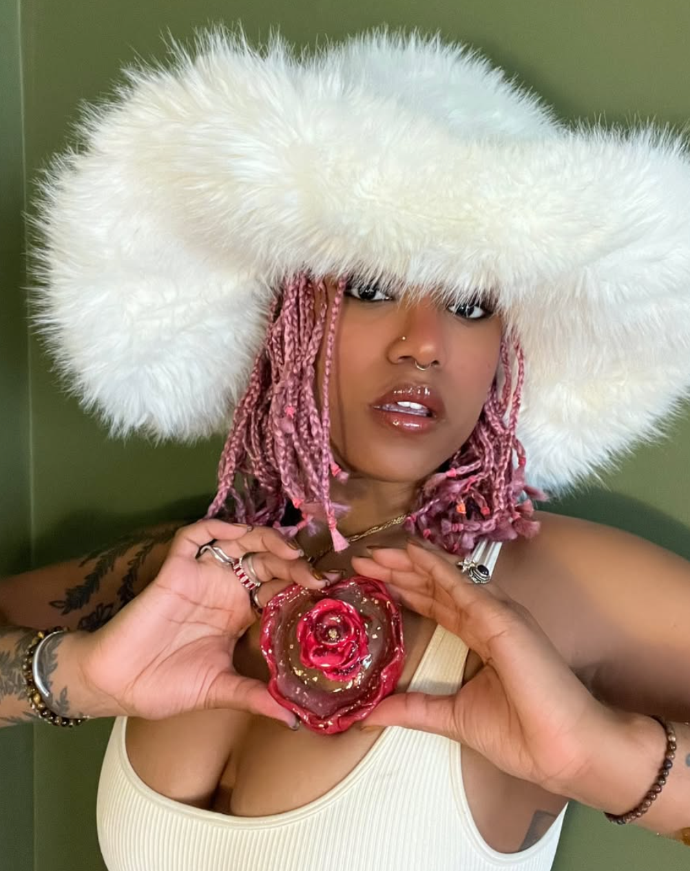 Woman in fluffy white hat and pink braids holds a rose-shaped pendant, showing a heart shape with hands.