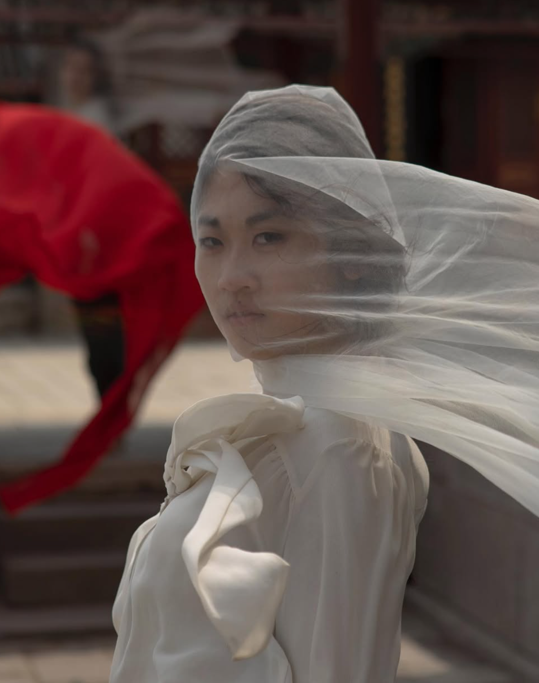 Woman in white veil gazes at camera; red flag in background; outdoor setting.