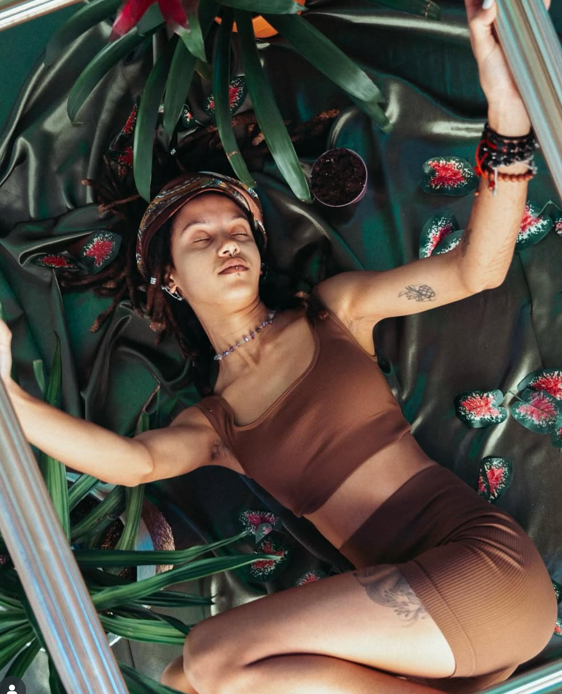 Woman in brown outfit reclines amidst green fabric and plants, arms outstretched.