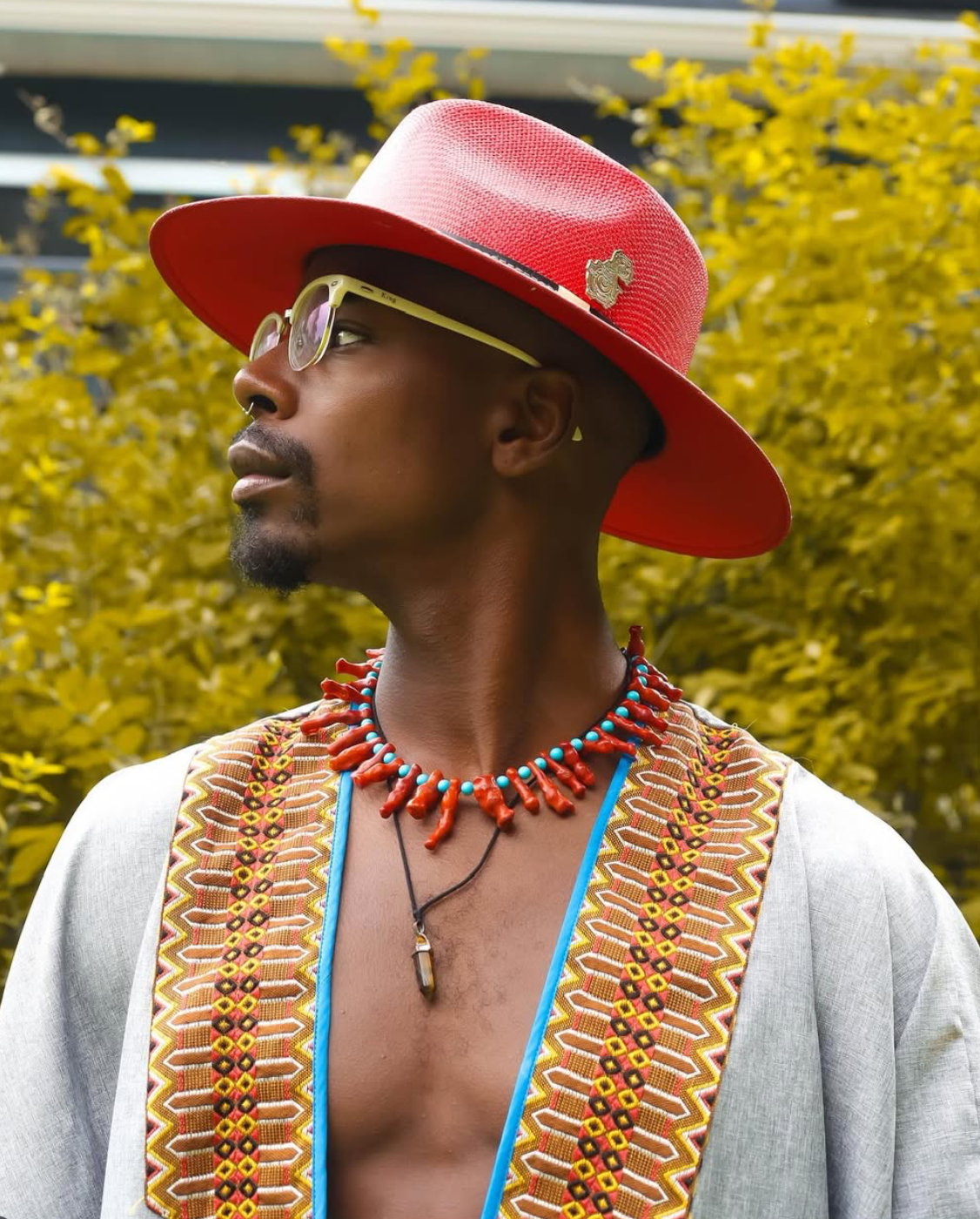 Man wearing red hat, glasses, colorful jewelry, and patterned shirt, set outdoors with yellow foliage.