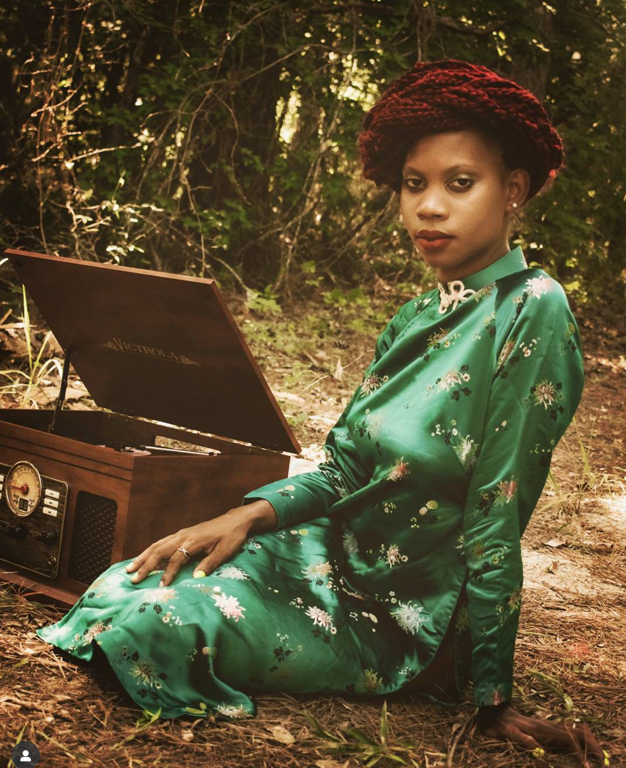 Woman in green dress and red hat sits by open vintage record player in woods.