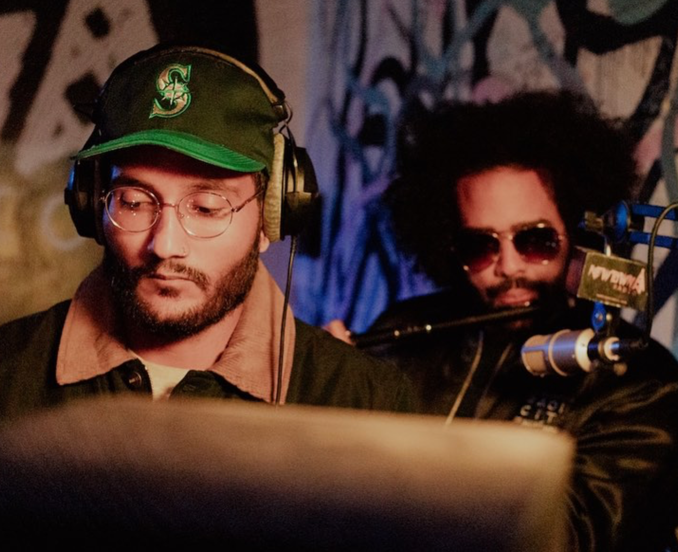 Two men at a radio station: one wearing a green baseball cap, the other with sunglasses, both near a microphone.