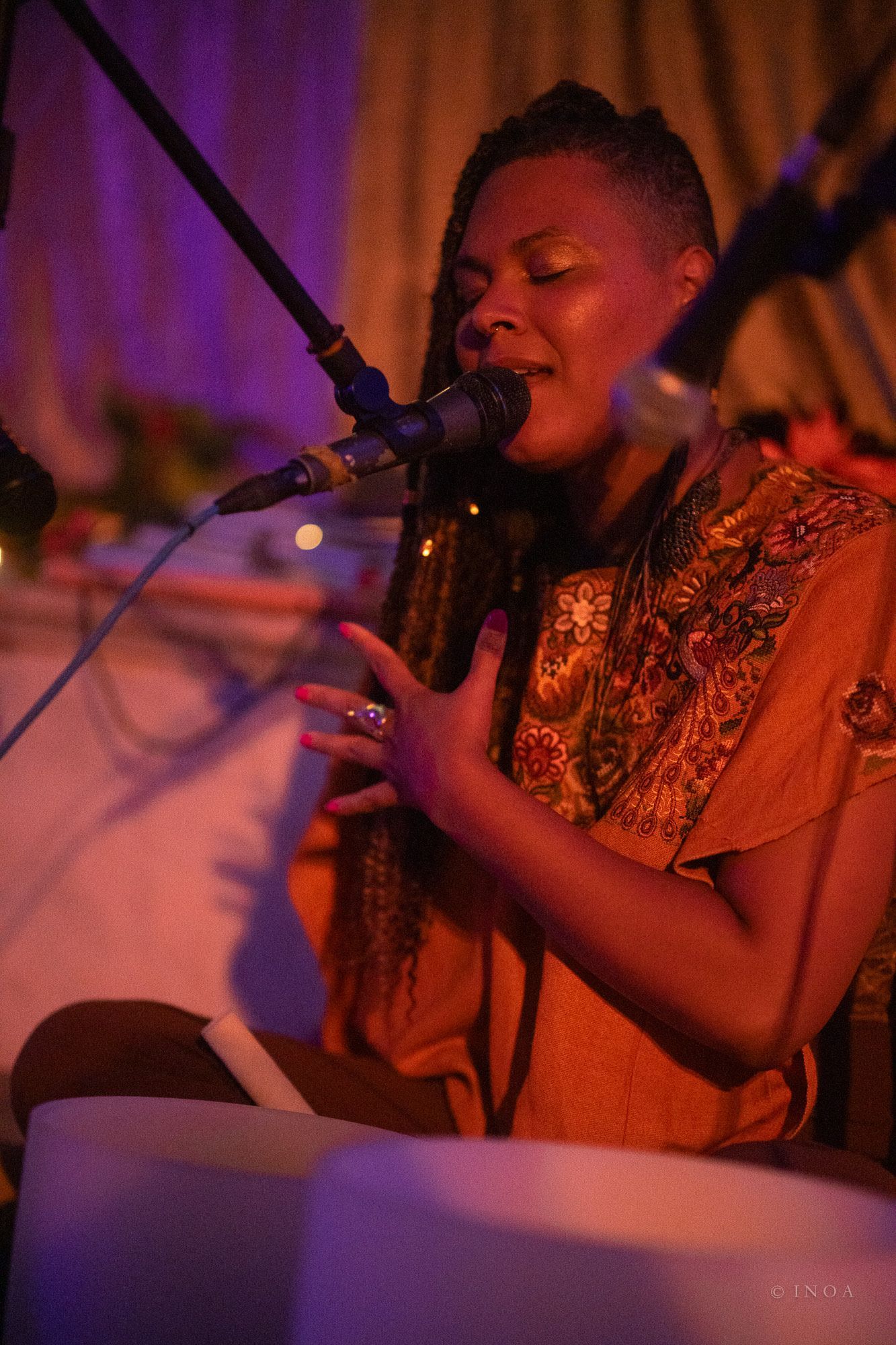 Woman singing, hands on chest, near microphones. Wearing gold top, in dim setting with singing bowls.