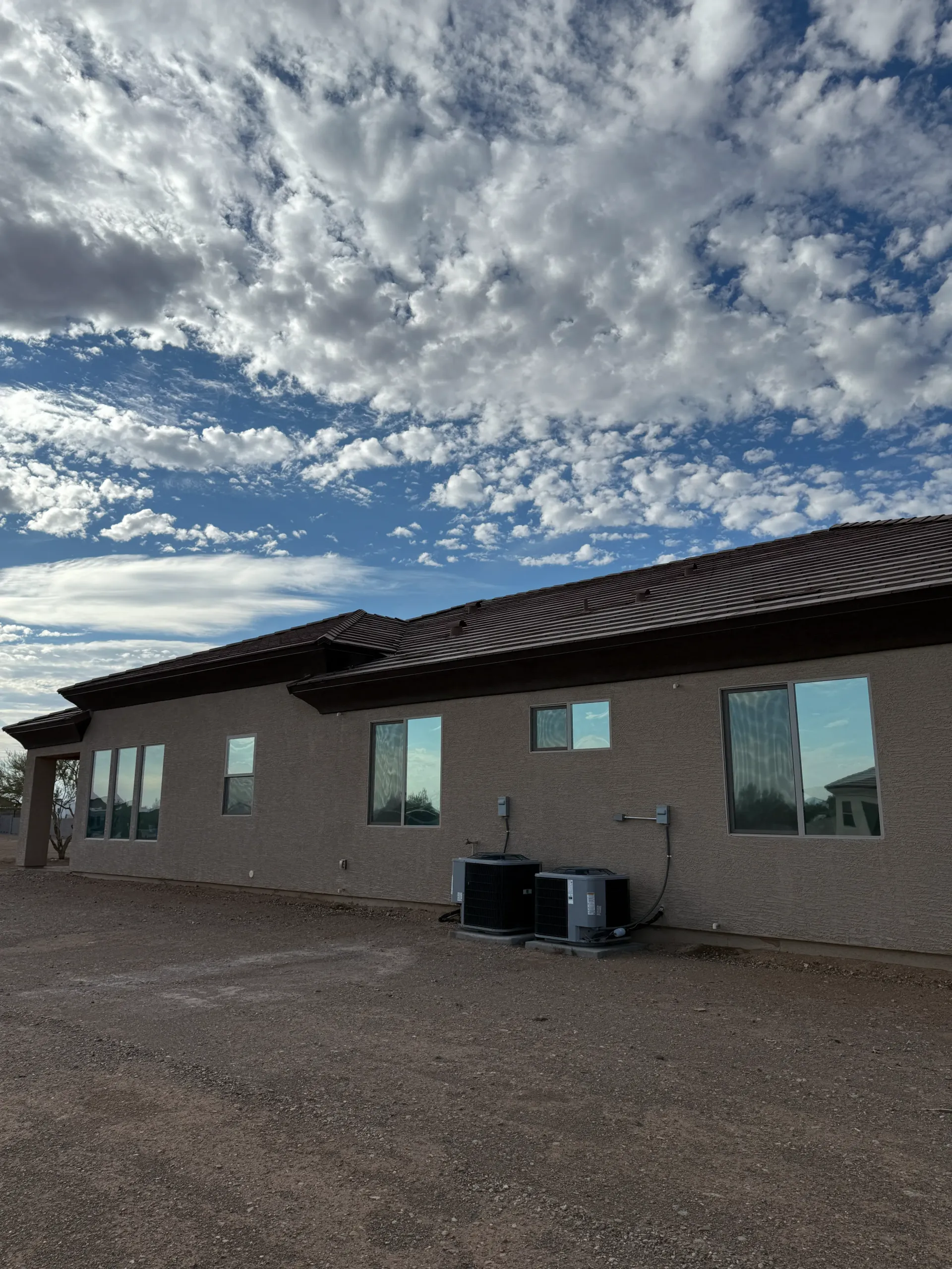 Tan stucco house with dark roof, windows reflecting the cloudy sky. Air conditioning units are visible.