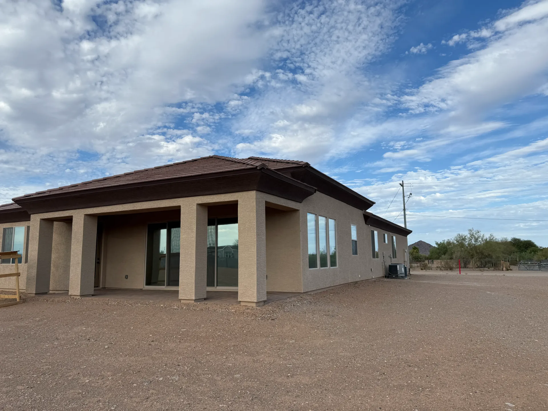 Tan stucco house with a covered patio, windows, and a gravel yard under a cloudy sky.