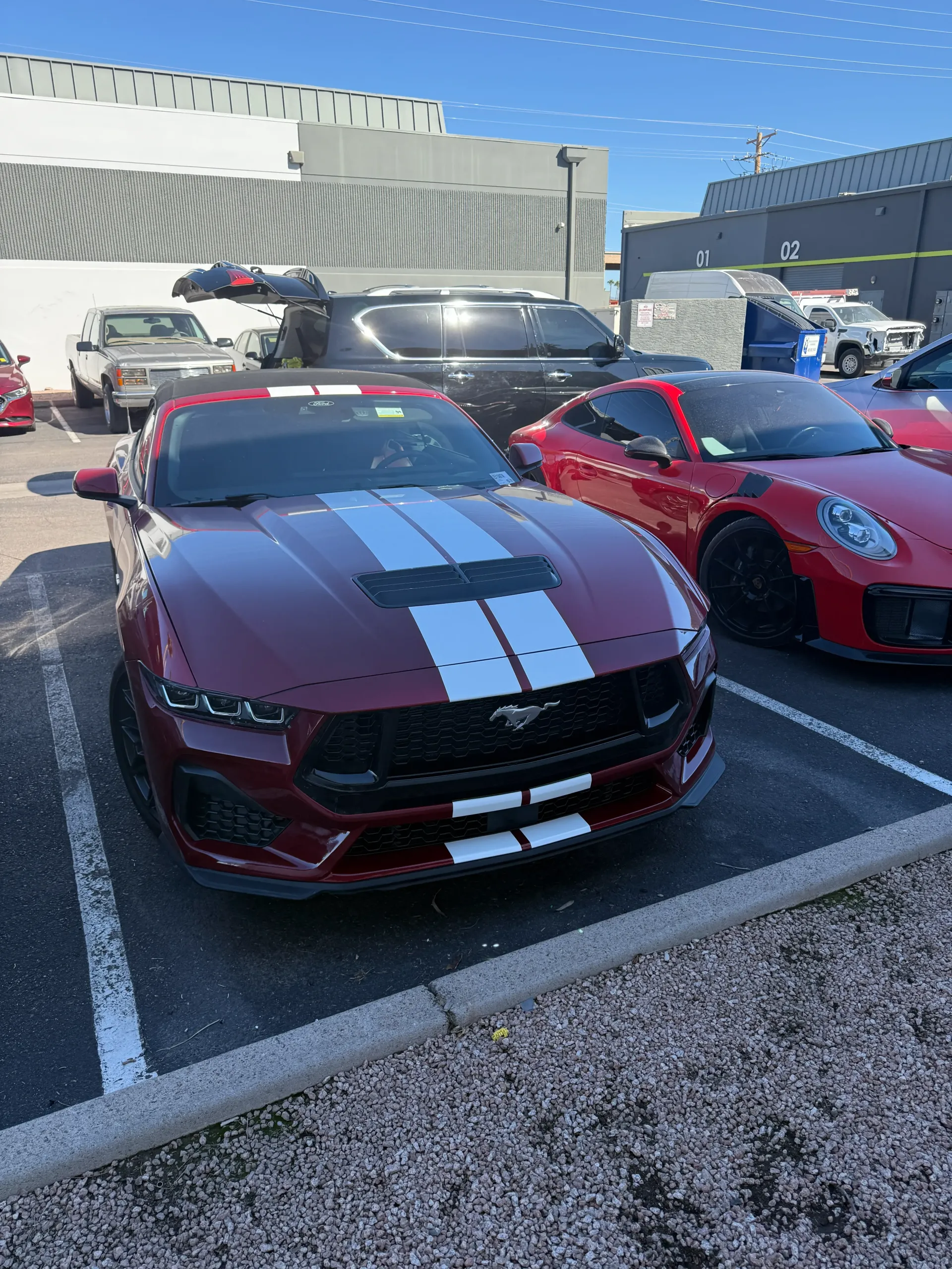 Red Ford Mustang with white stripes parked next to a red Porsche, in a parking lot.