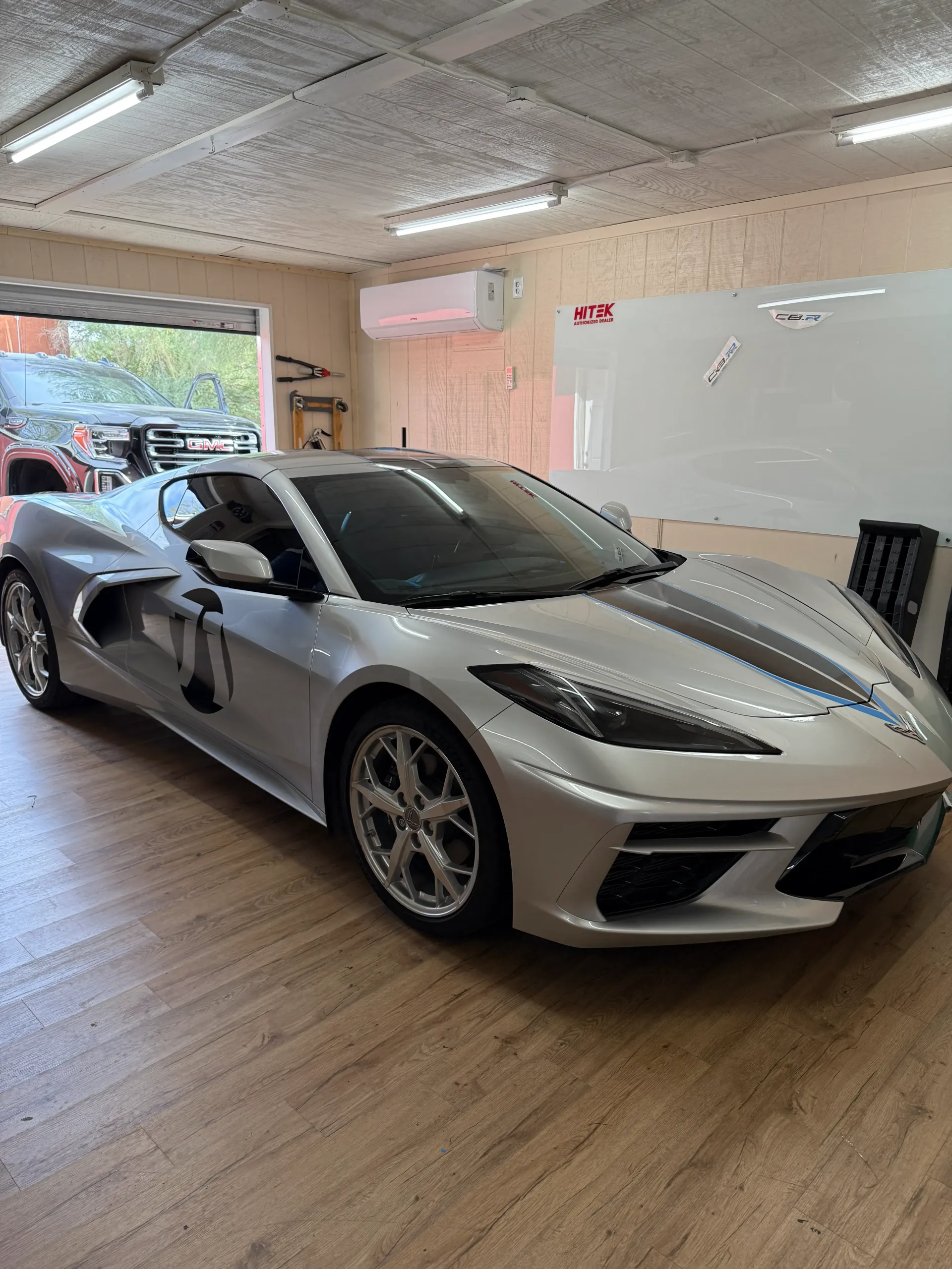 Silver Corvette sports car inside a garage, with a black stripe down the middle, on wood flooring.