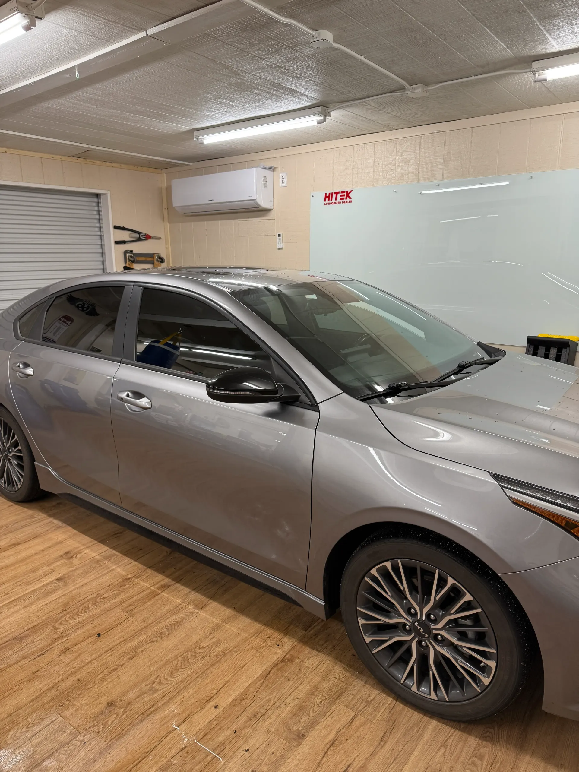 Silver Kia sedan parked indoors with a white wall and an air conditioning unit.