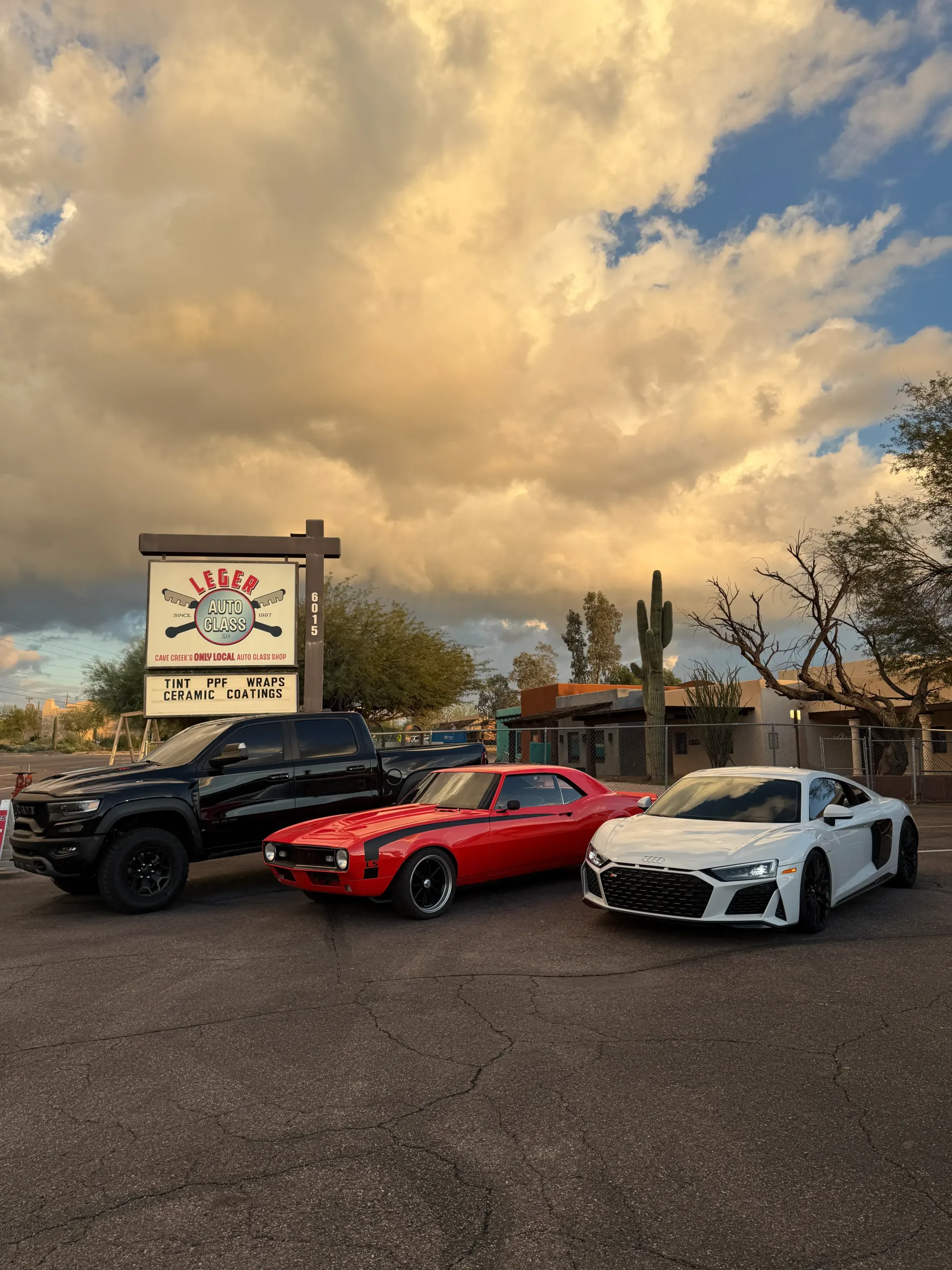 Three vehicles parked outside a sign. A black truck, red classic car, and white sports car under a cloudy sky.