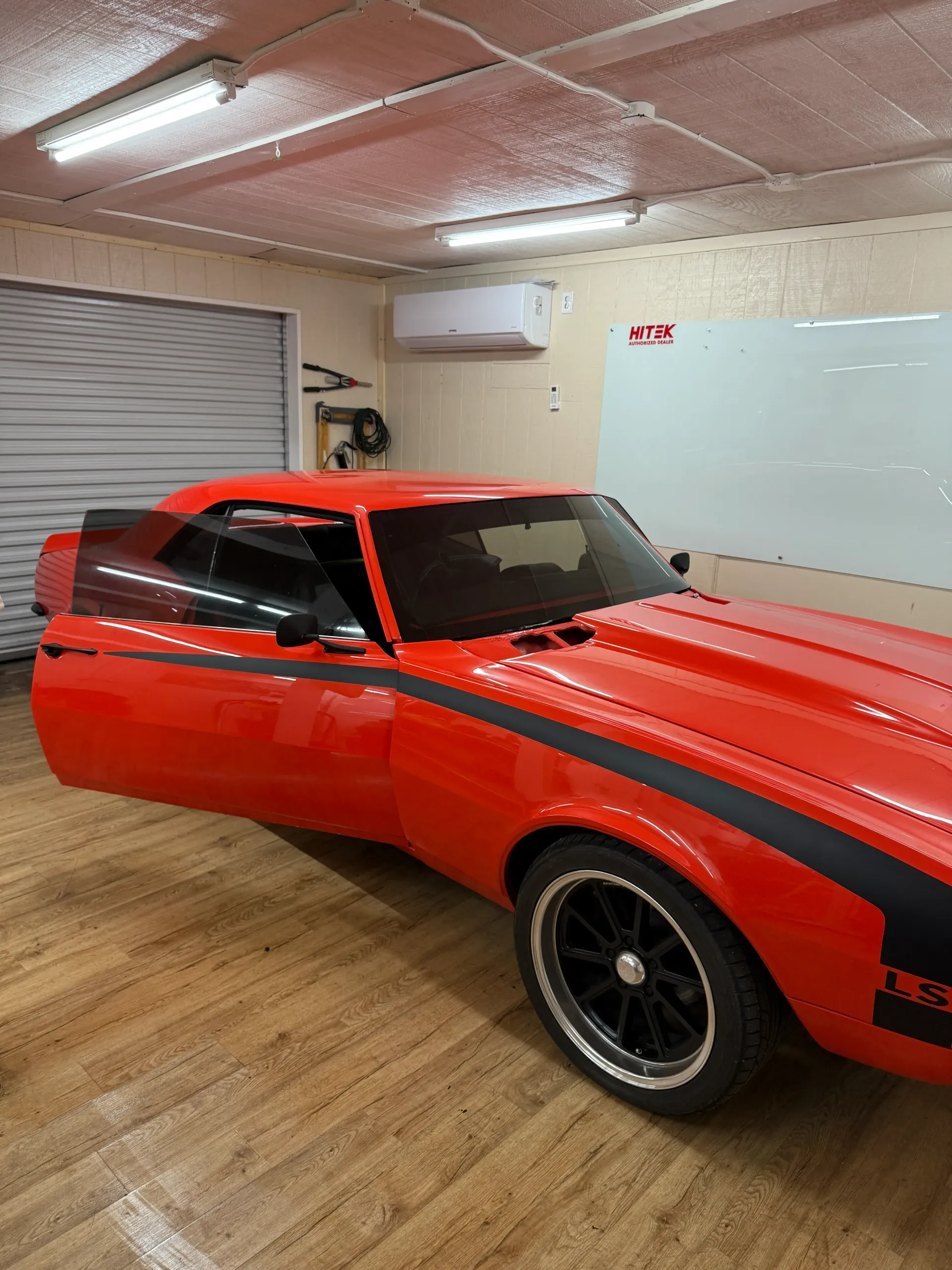 Bright orange classic car with black racing stripes, door open, parked indoors.