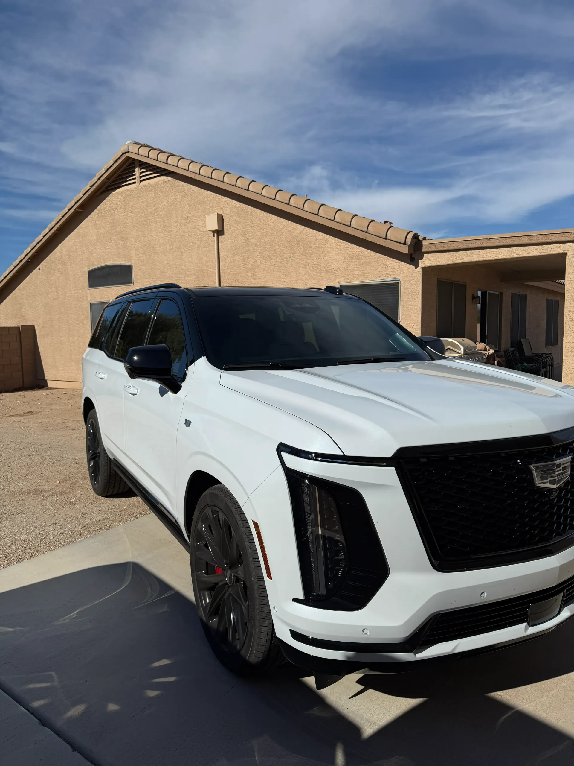 White Cadillac SUV parked in front of a tan house on a sunny day. Black roof, grill, and wheels.