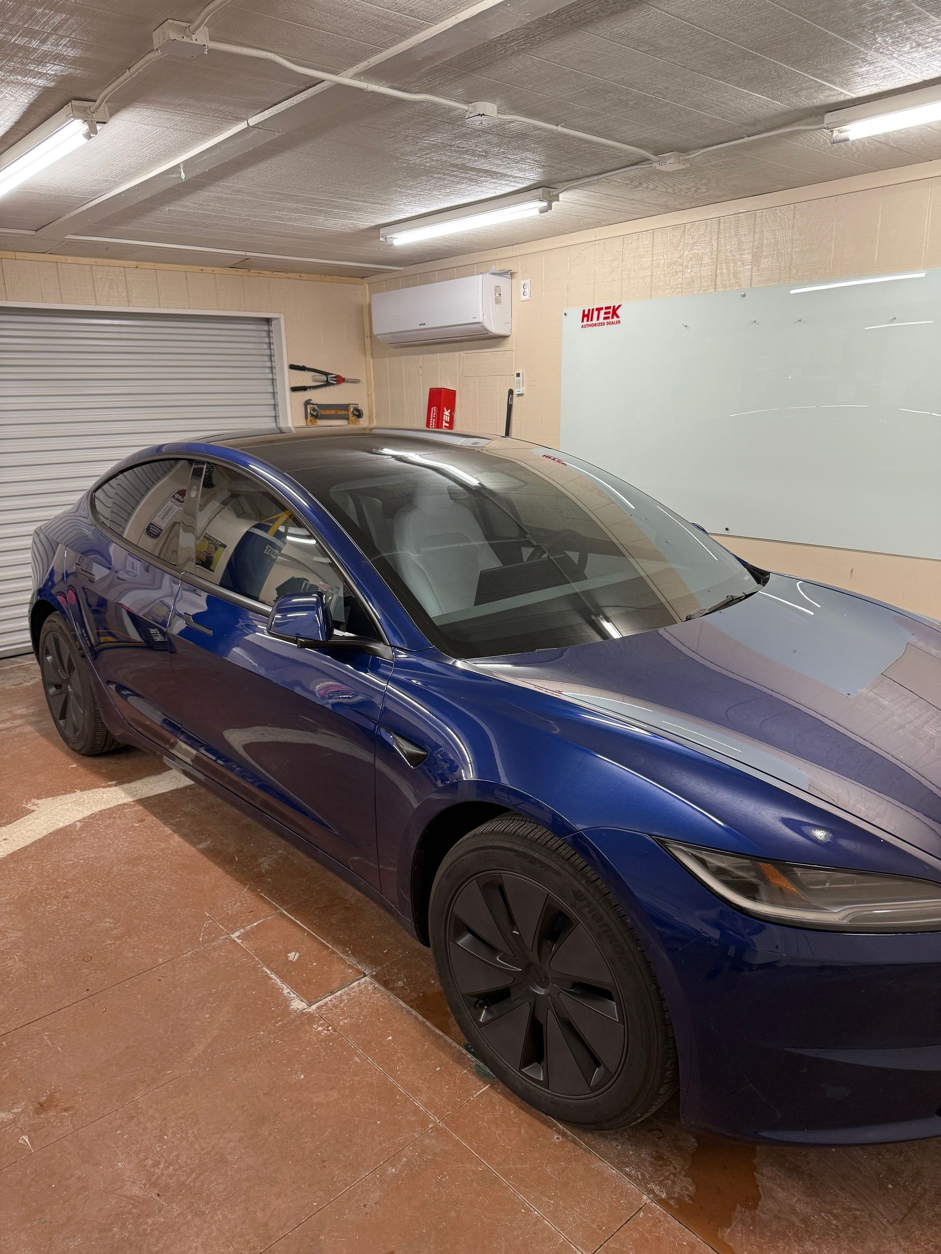 Blue Tesla car parked in a garage with a roll-up door and a white board.