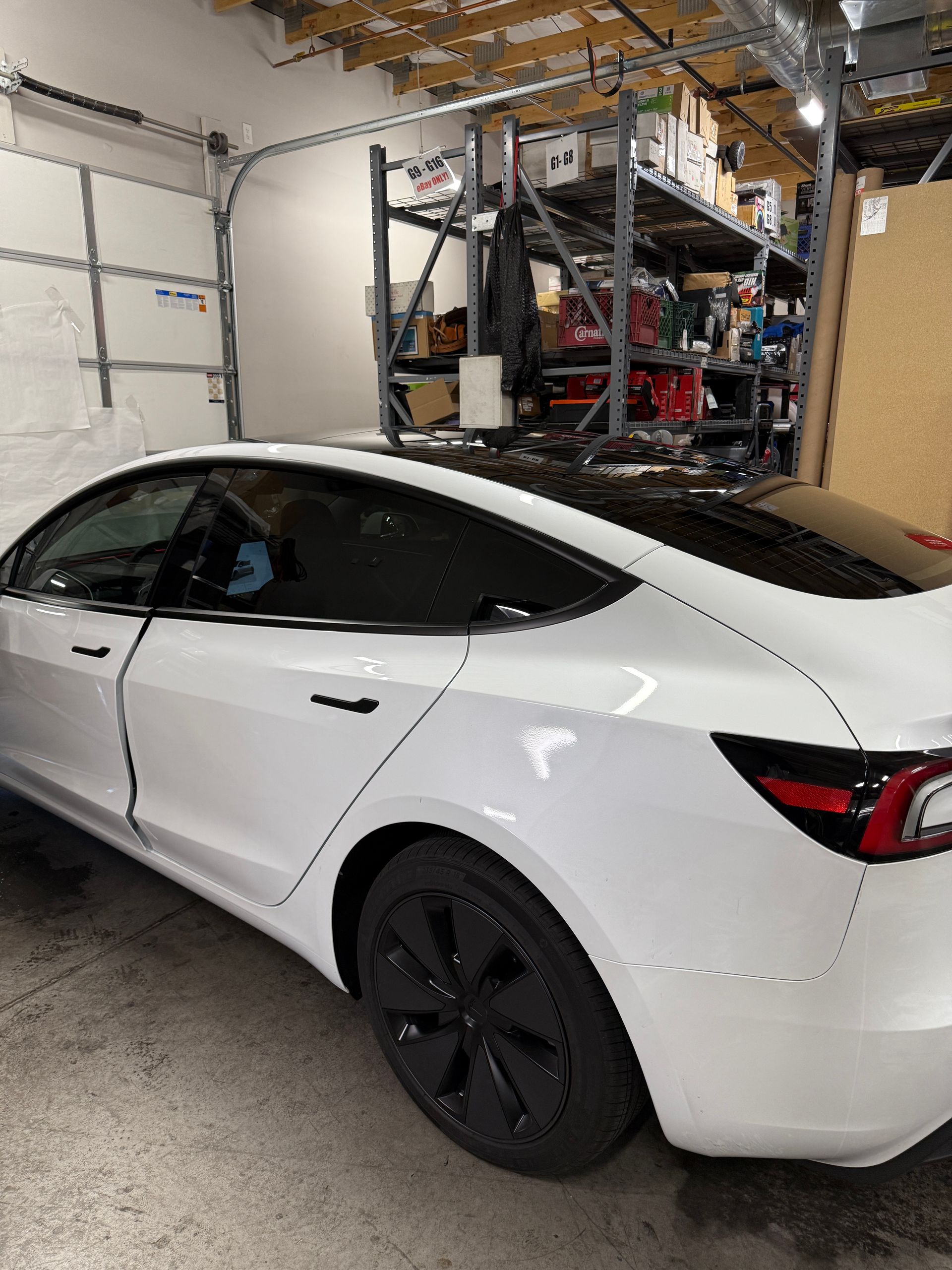 White Tesla Model 3 with black accents in a garage, next to a garage door and shelving.