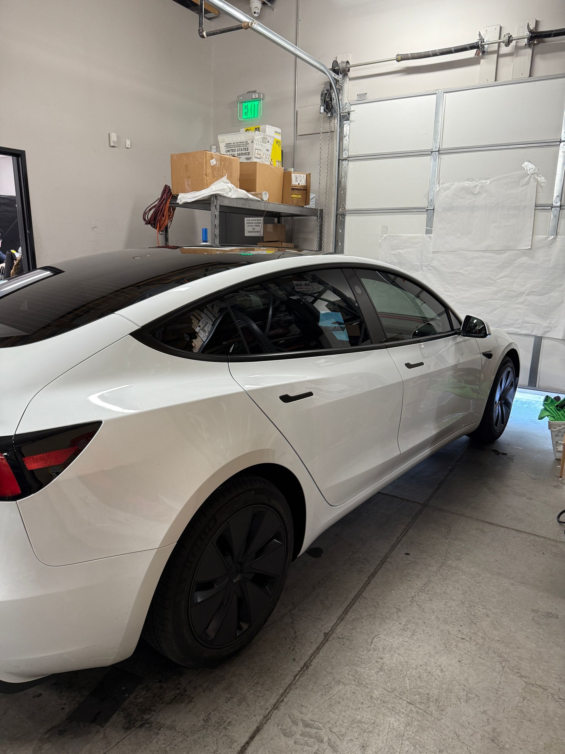 White Tesla car in a garage with a partially open door. Black wheels and trim. Boxes on a shelf in background.
