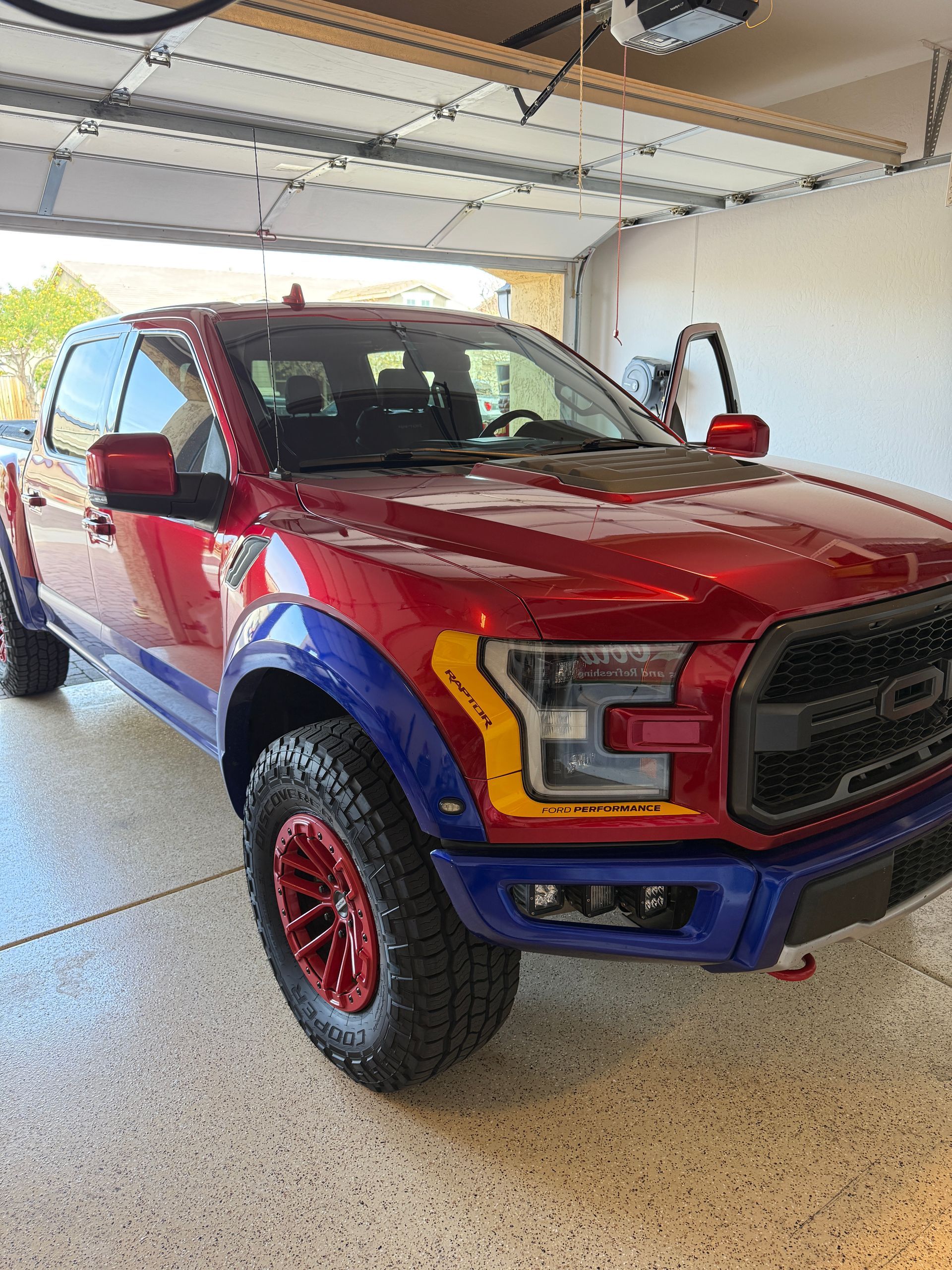Red, blue, and yellow Ford Raptor truck parked in a garage with open door, red wheels.