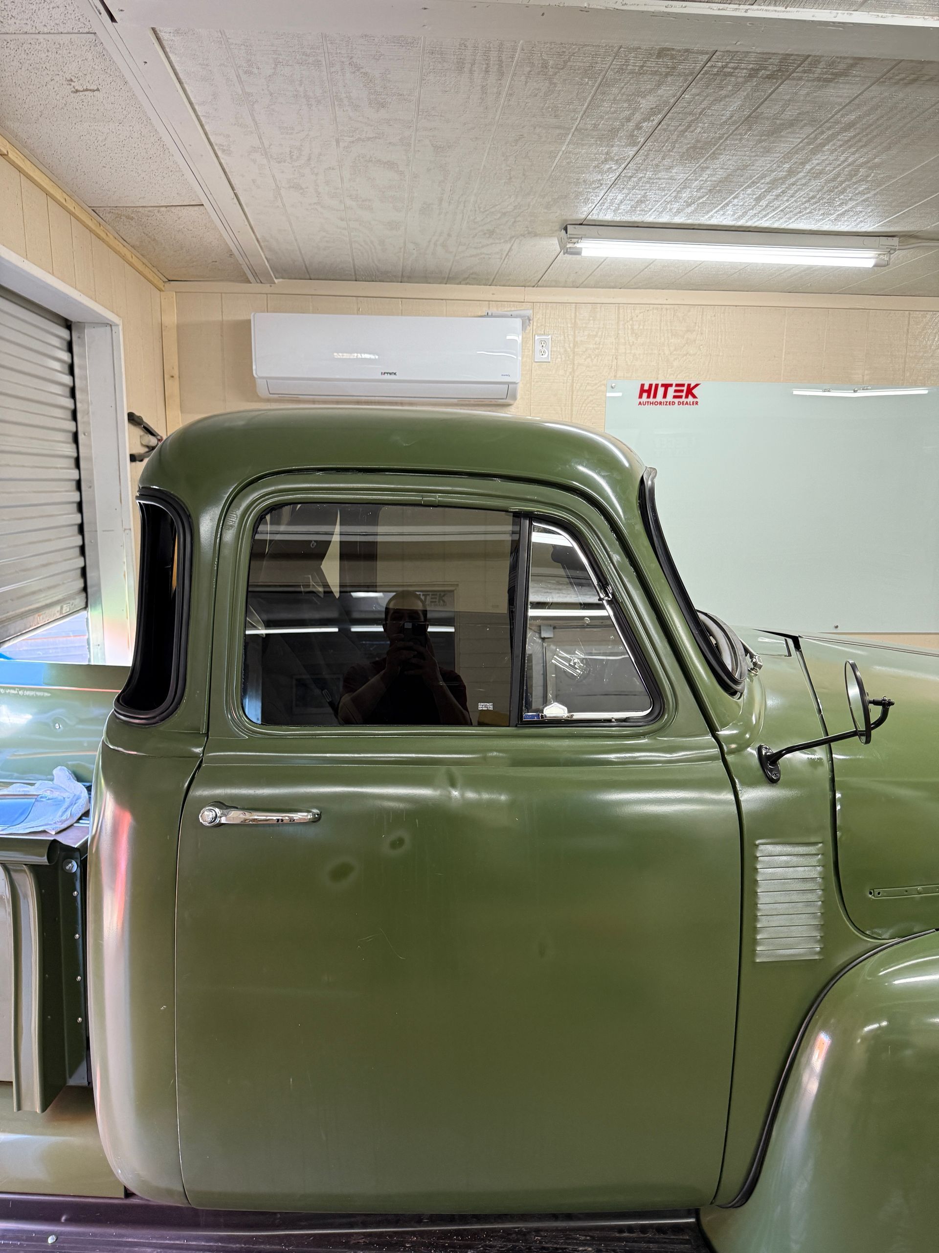Green classic truck cab in a workshop, door visible, window reflecting the room, air conditioning unit in the background.