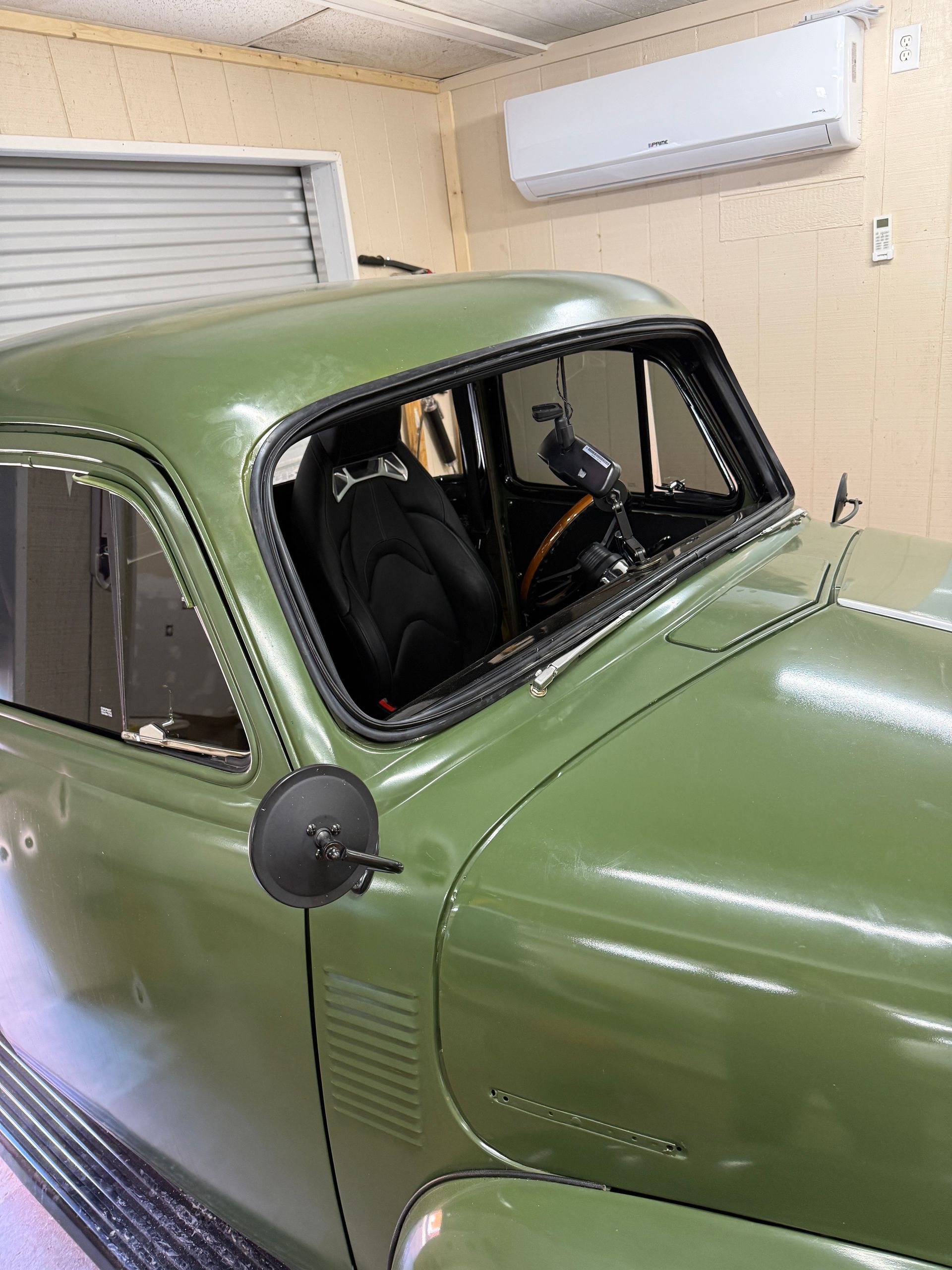 Olive green classic car parked in a garage with a black interior. Side mirror and windshield are visible.