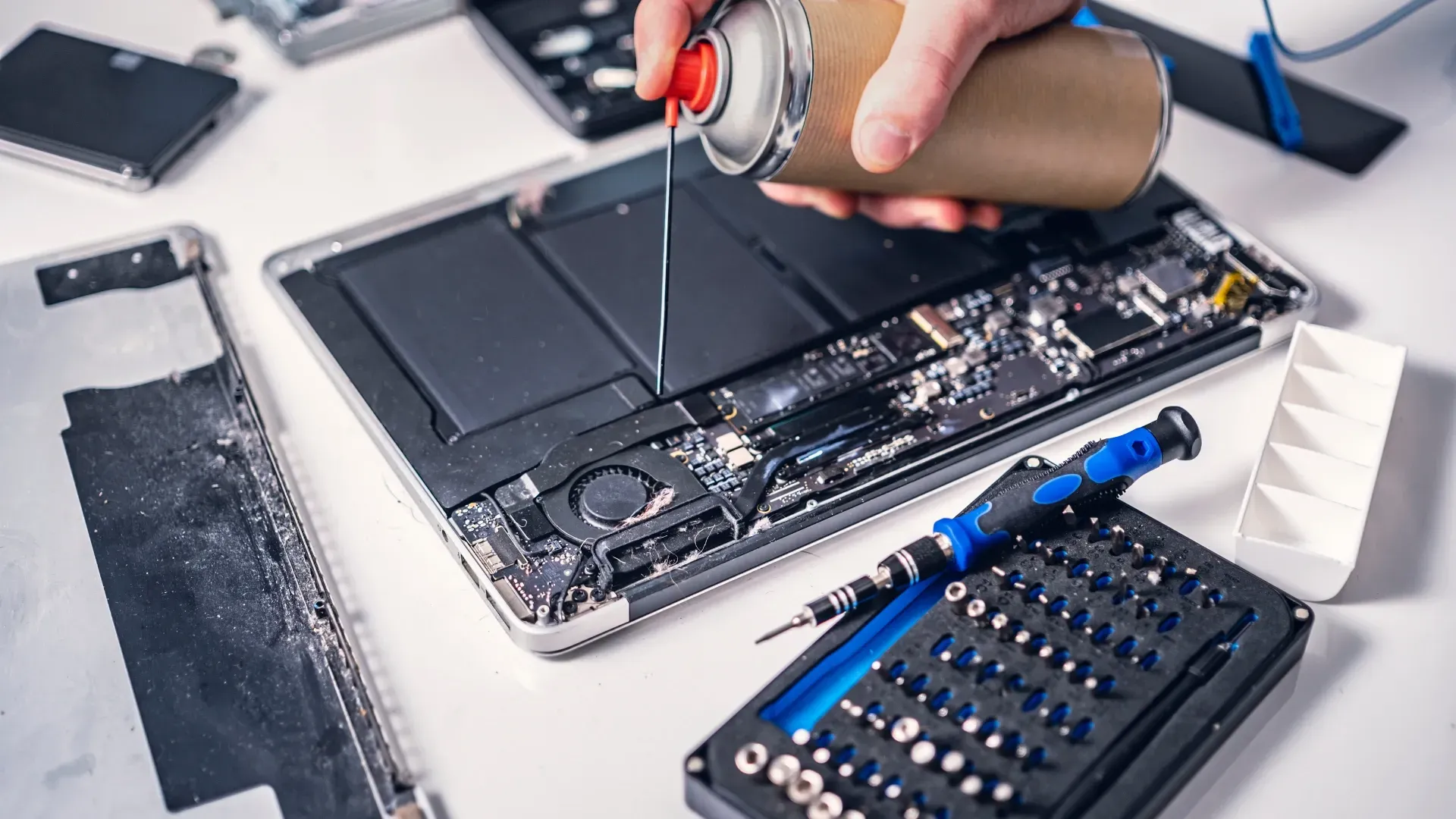 technician blowing dust & debris from laptop with case opened during repairs using air duster with professional repair tools on repair desk