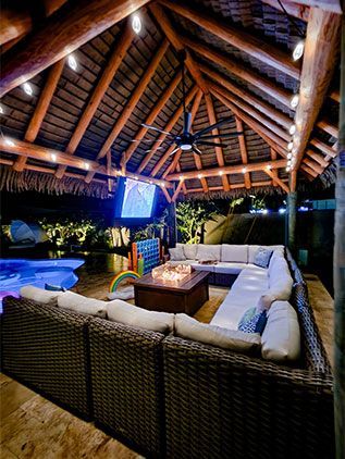 Outdoor lounge with thatched roof, sectional sofa, fire pit, and pool at night. Outdoor lounge with thatched roof, sectional sofa, fire pit, and pool at night.