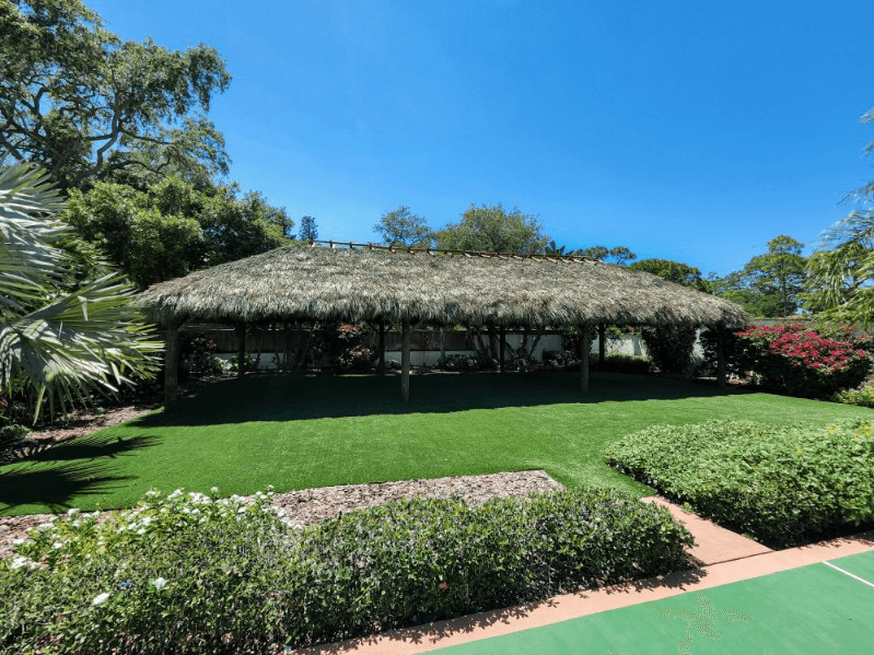 A thatched-roof structure stands over green grass, surrounded by greenery and a blue sky.