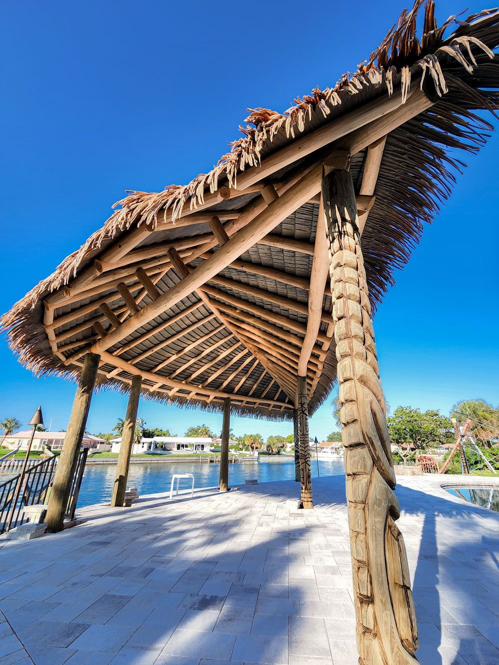 Tiki-style wooden pavilion with thatched roof, overlooking water under a bright blue sky.