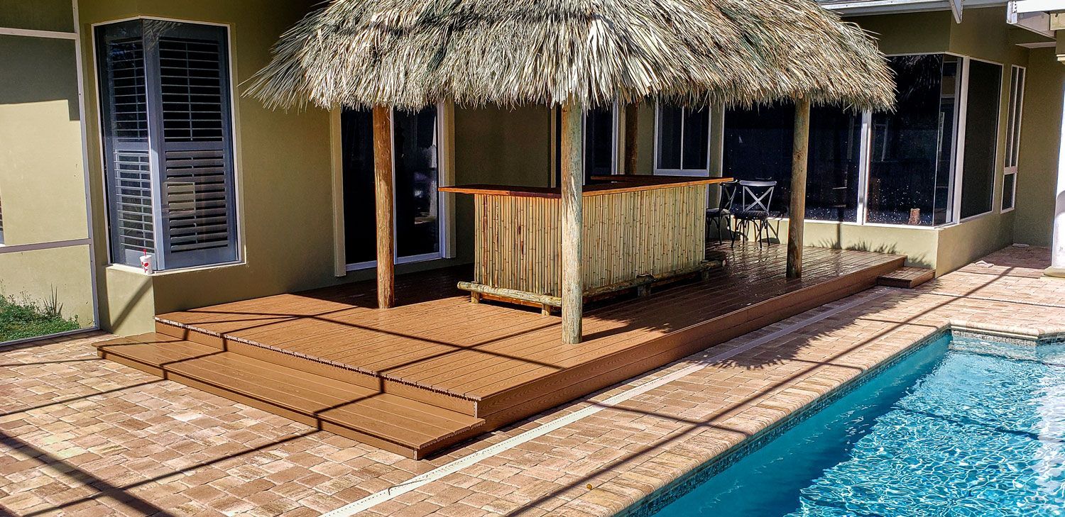 Tiki bar with thatched roof next to a swimming pool on a wooden deck.
