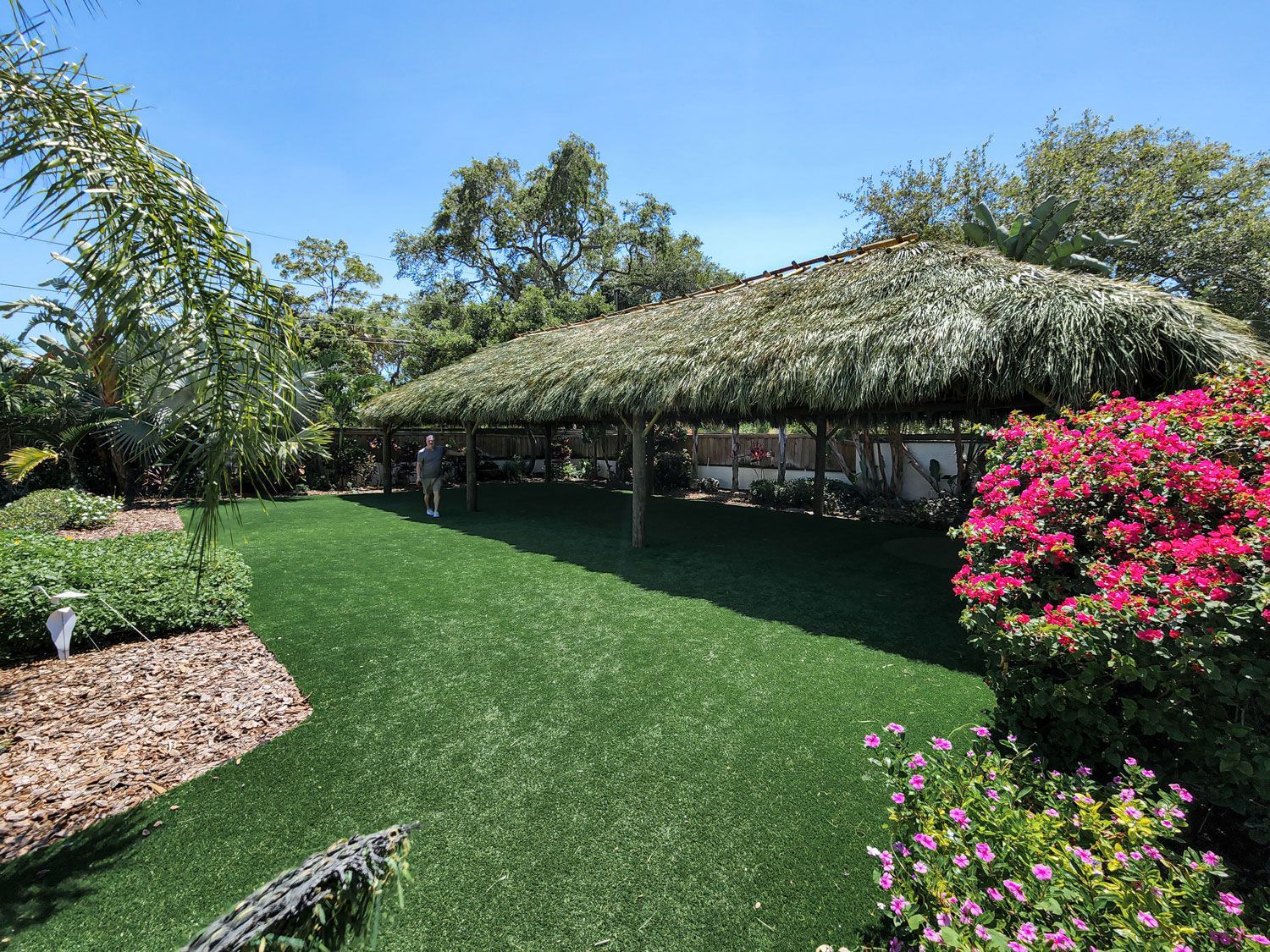 Long, grassy shelter with a thatched roof in a garden, bright pink flowers on right, person standing in the shade.