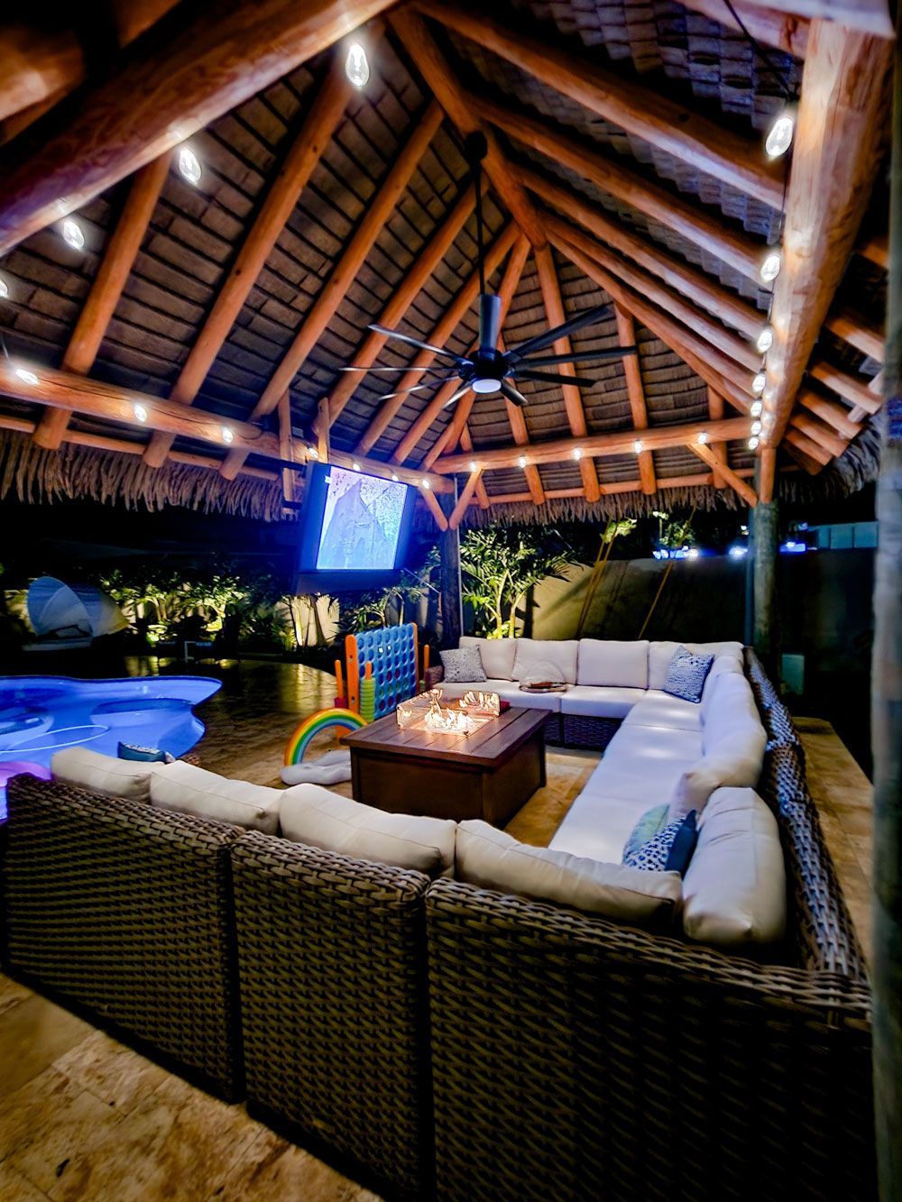 Outdoor patio with thatched roof, sectional sofa, fire pit, pool in background, string lights.