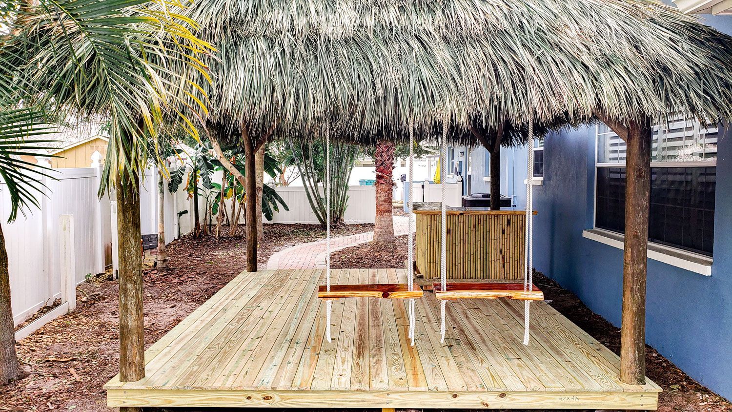 Wooden deck with a thatched roof, swings, and palm trees in a backyard.