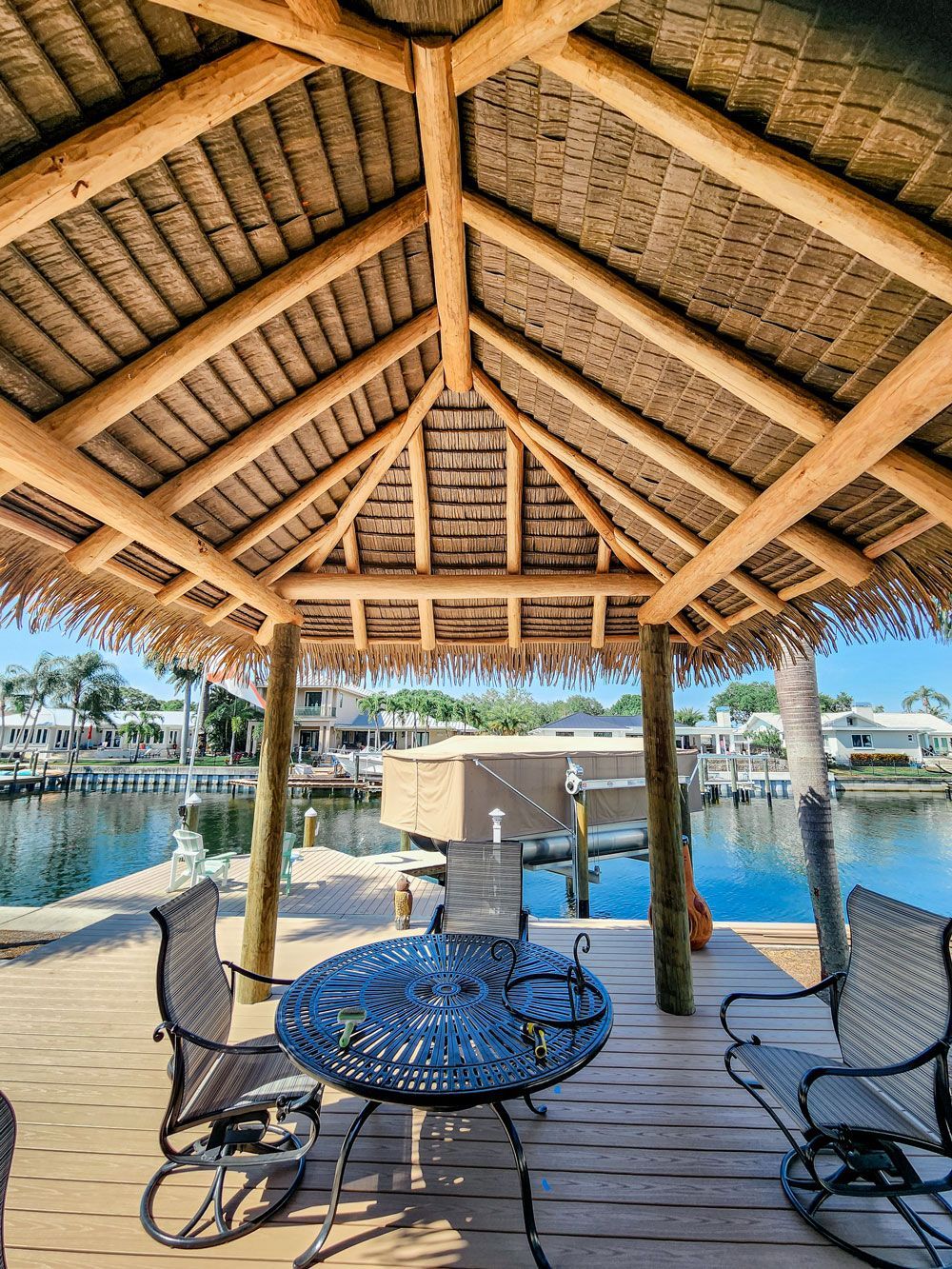 Gazebo over water with table, chairs, and houses in the background. Wooden beams support the thatched roof.