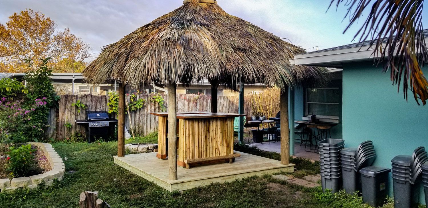 Tiki hut with a wooden bar in a backyard. The hut has a thatched roof, and is surrounded by green grass and a wooden fence.